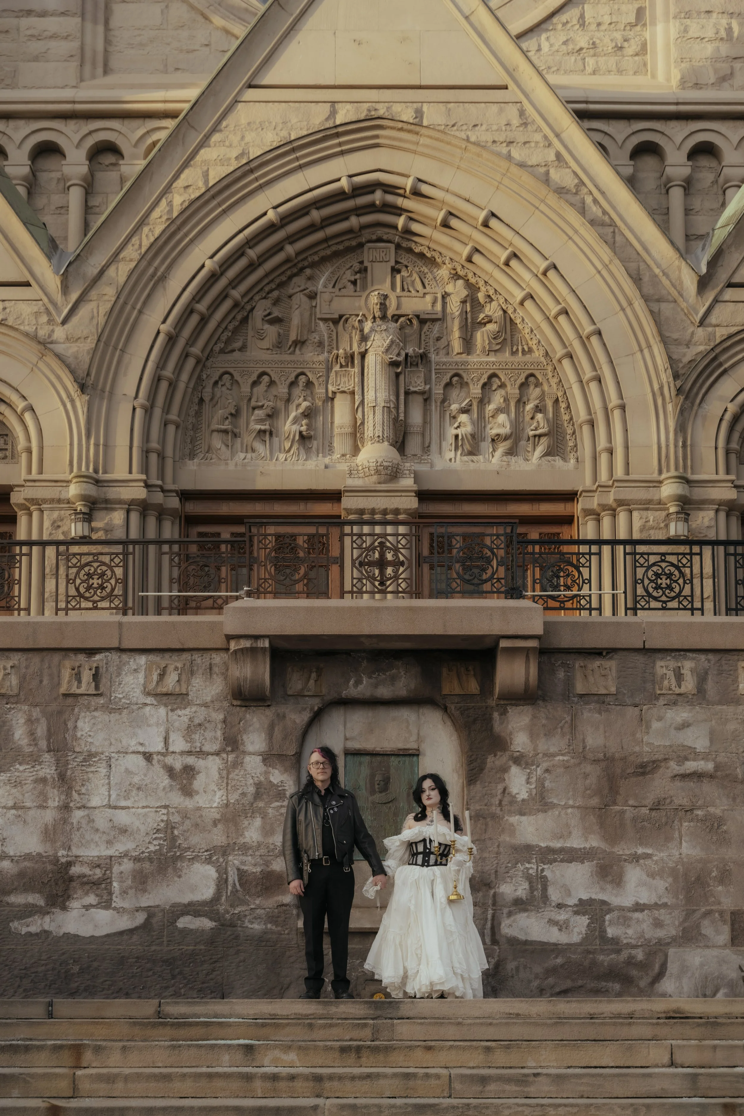 Two women stand hand in hand in front of a stone church with a large sculpted arch and religious figures. One woman is dressed in a black leather jacket and dark pants, the other in a white gown with a black corset and holding a candle and a goblet.
