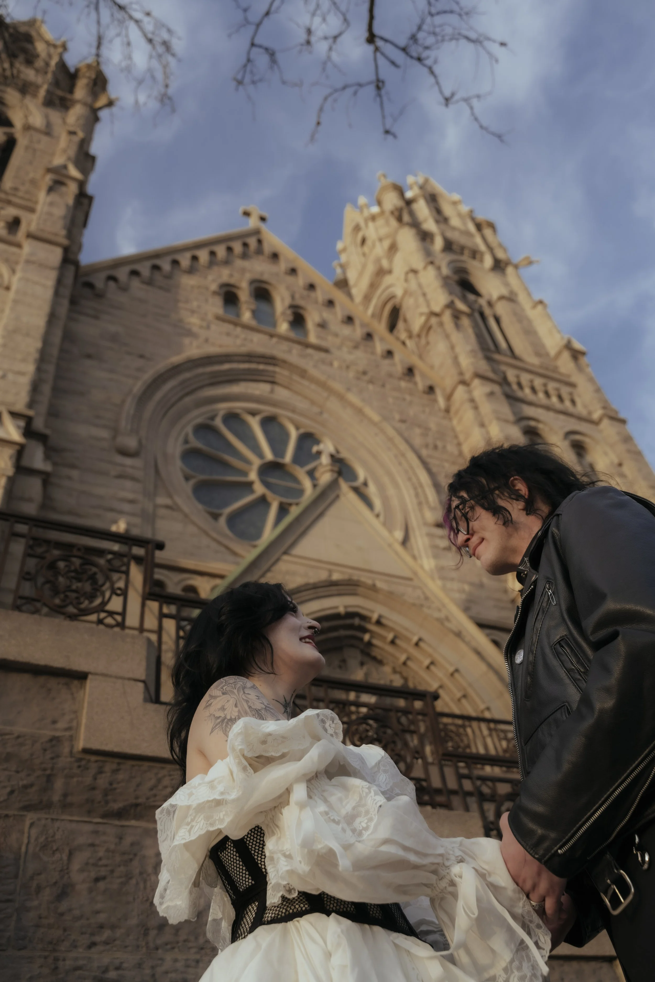 A couple standing in front of a historic church, smiling and holding hands, with the church's Gothic architecture and a large clock in the background.