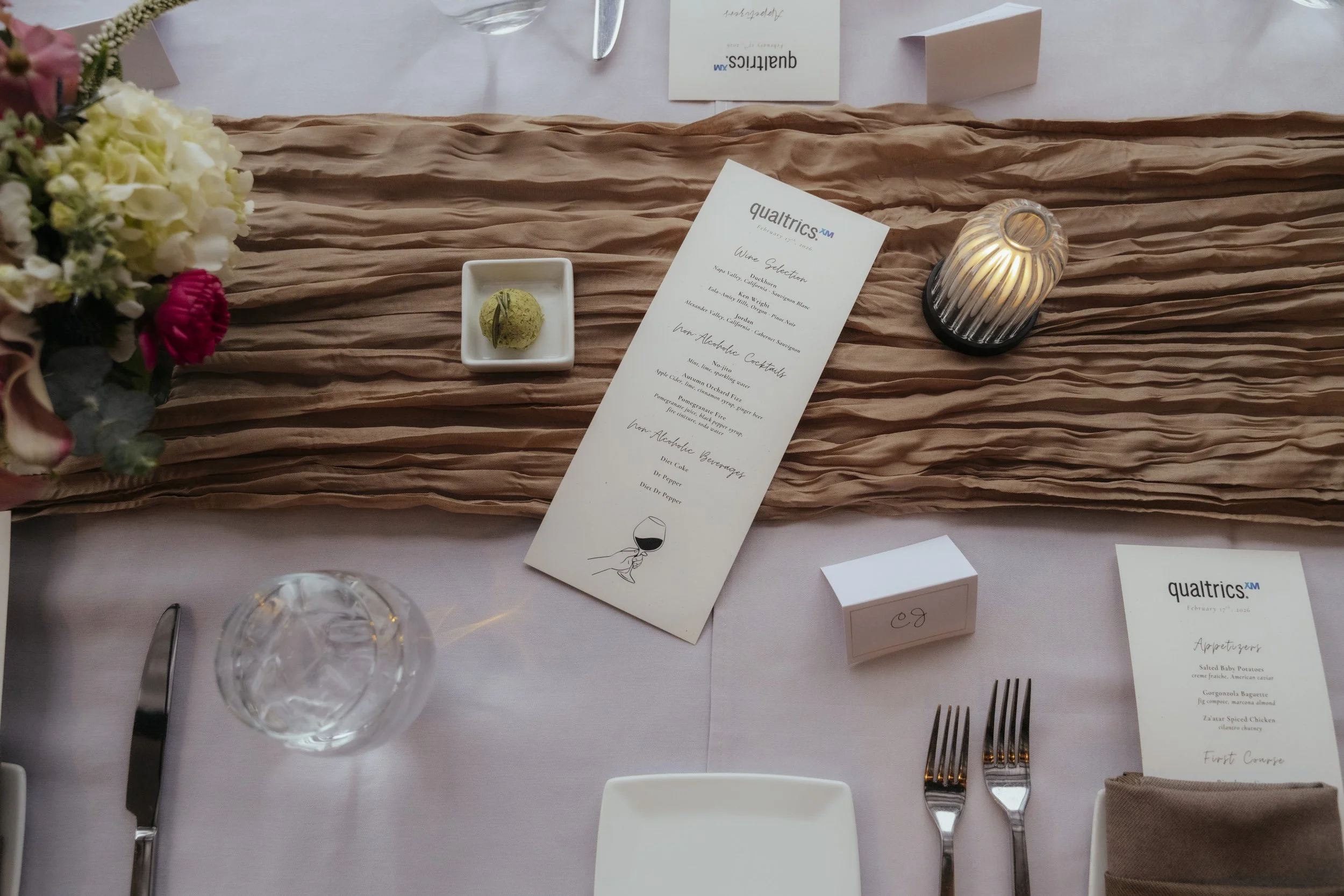 Elegant table setting with a floral centerpiece, a crinkled peach-colored table runner, printed menus, a green dessert, a wine glass, and lit candle for Qualtrics