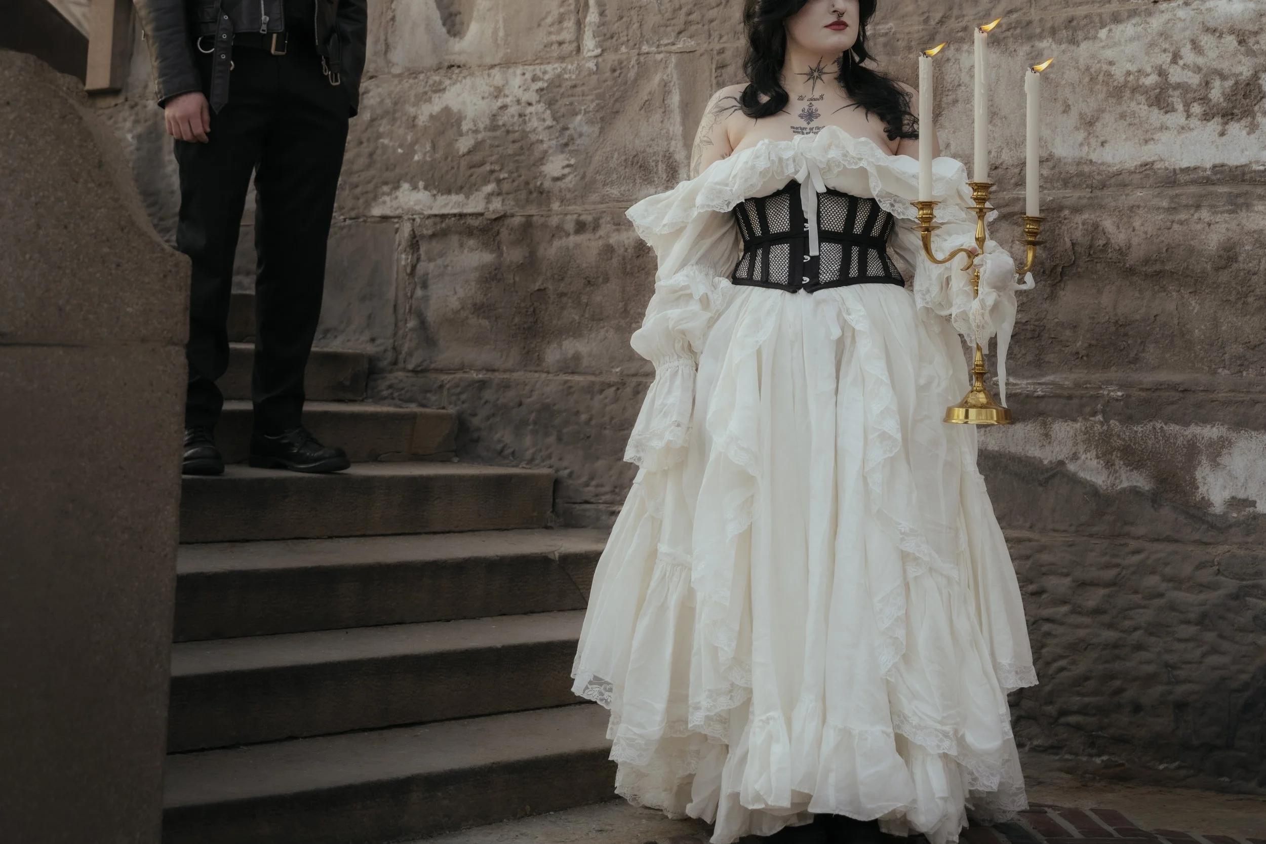 A woman in a vintage, off-the-shoulder cream-colored dress with lace and ruffles, wearing a black corset, holding a gold candlestick with three lit white candles, standing on stone stairs against a brick wall. A man in black pants and shoes is partia
