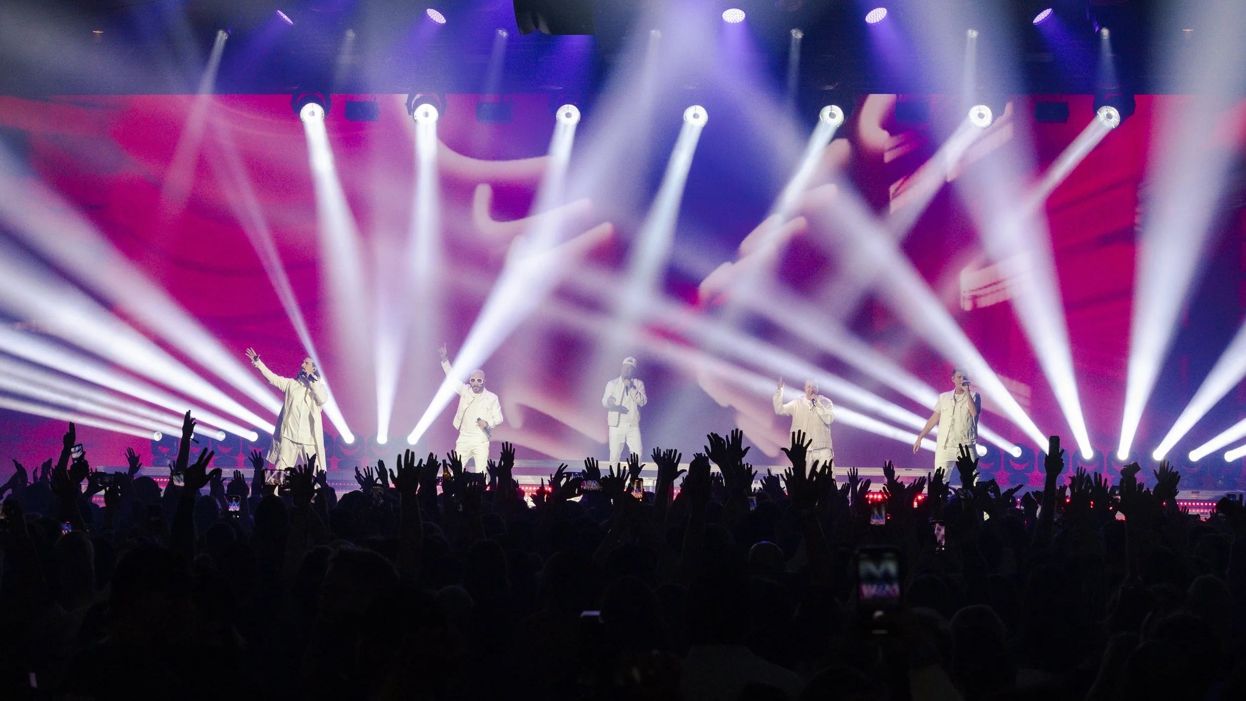 A concert stage with five performers (backstreet boys) dressed in white, surrounded by bright white and purple lighting, with an audience raising their hands in front.