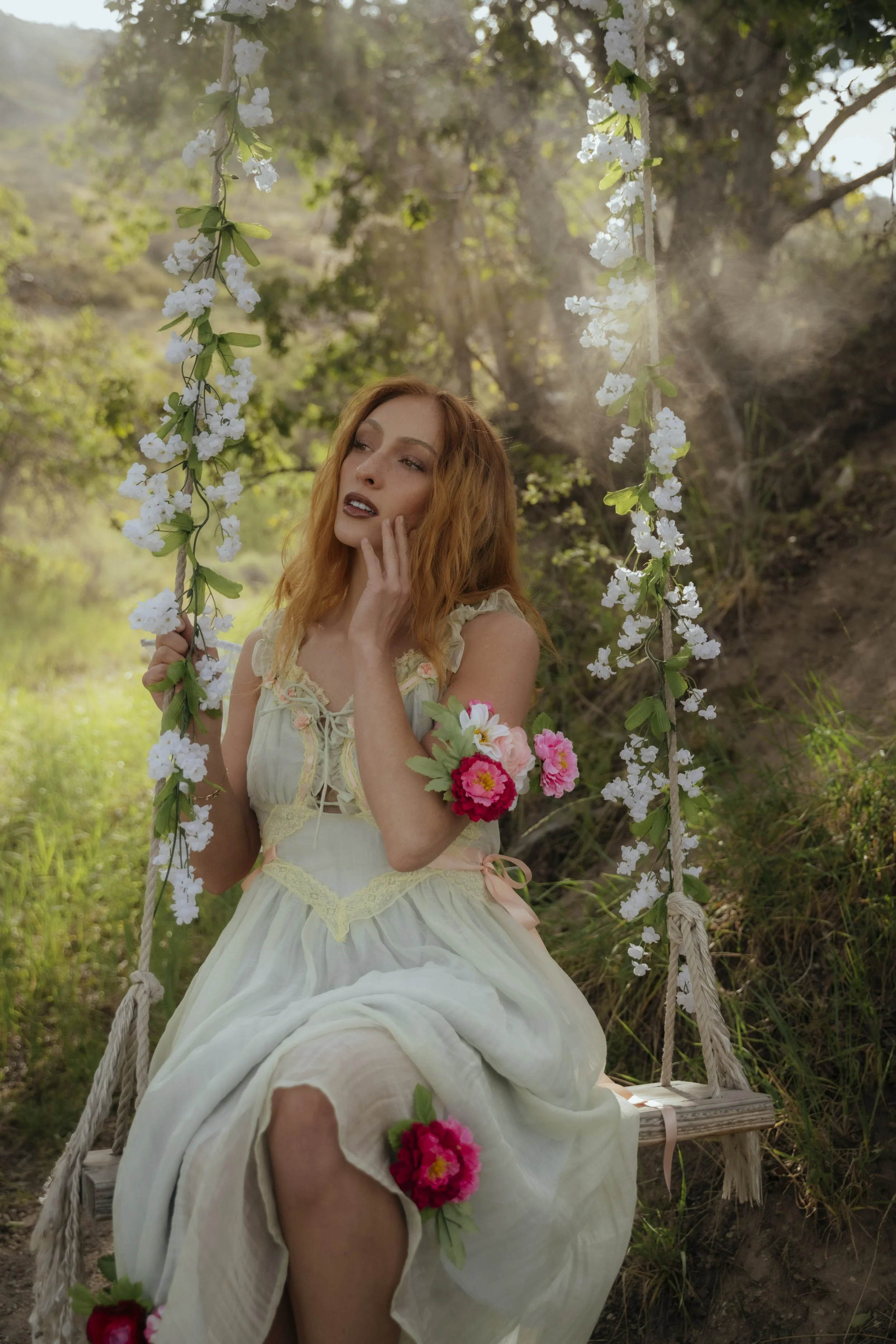 A young woman with red hair and fair skin sitting on a wooden swing adorned with white flowers, outdoors in a lush green setting. She is wearing a cream-colored dress with lace details and pink and red flowers on her wrist and around her waist, with 