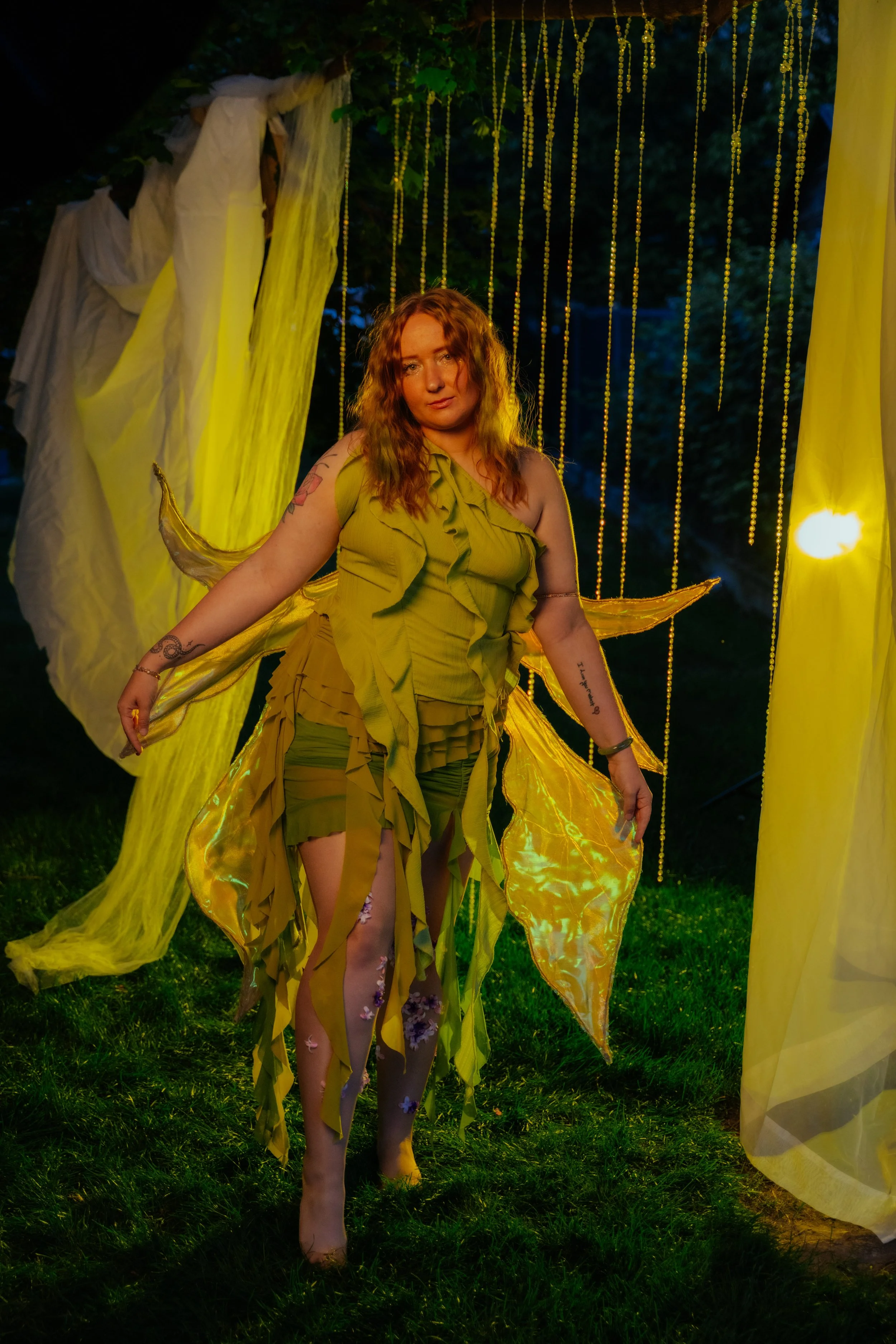 A woman with red hair dressed in yellow fairy-like costume, standing outdoors at night in front of yellow and white fabric decorations. She has tattoos on her arms and floral tights, with glowing yellow wings behind her.