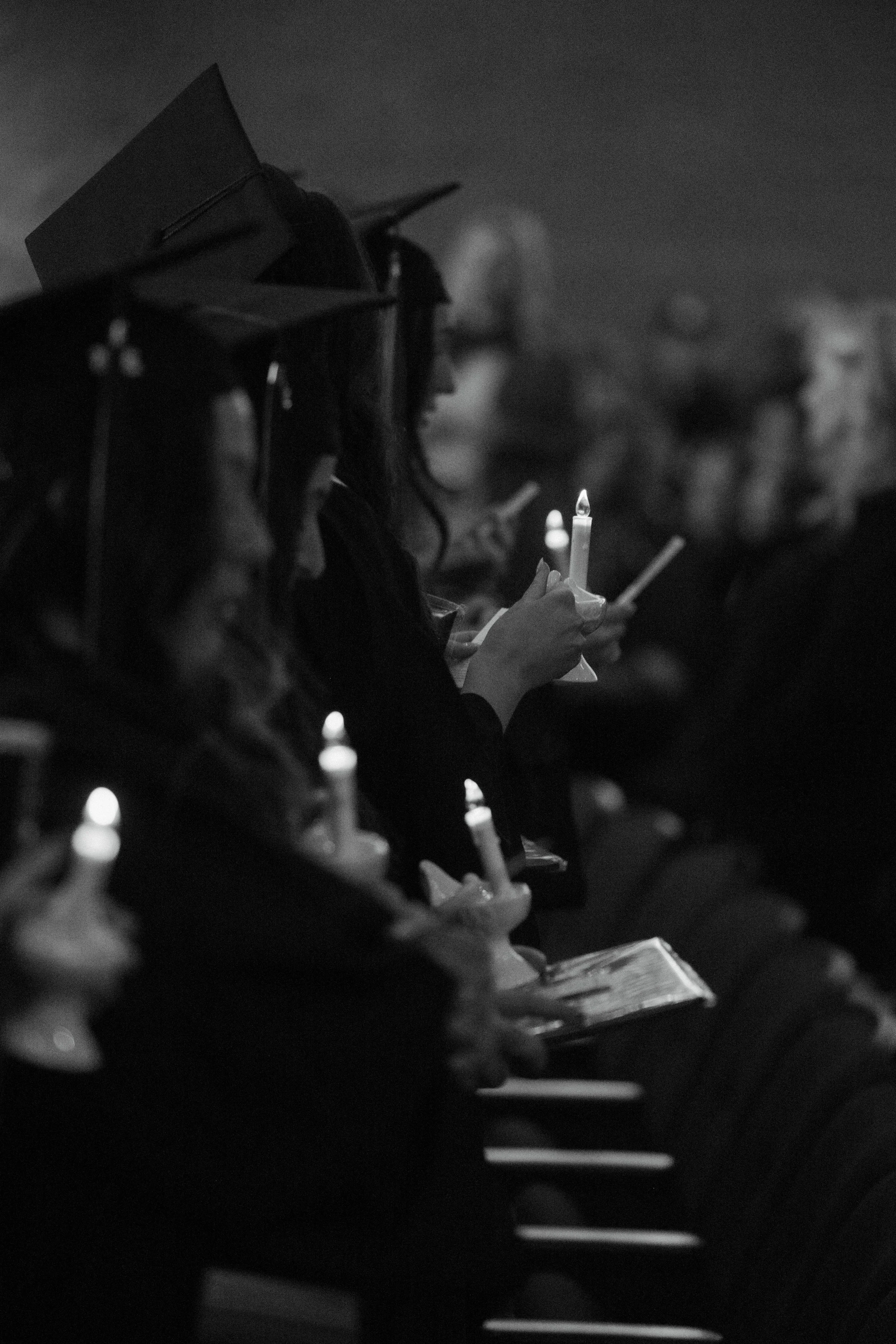 Graduates in caps and gowns holding candles during a ceremony, seated in a row in a dimly lit setting.
