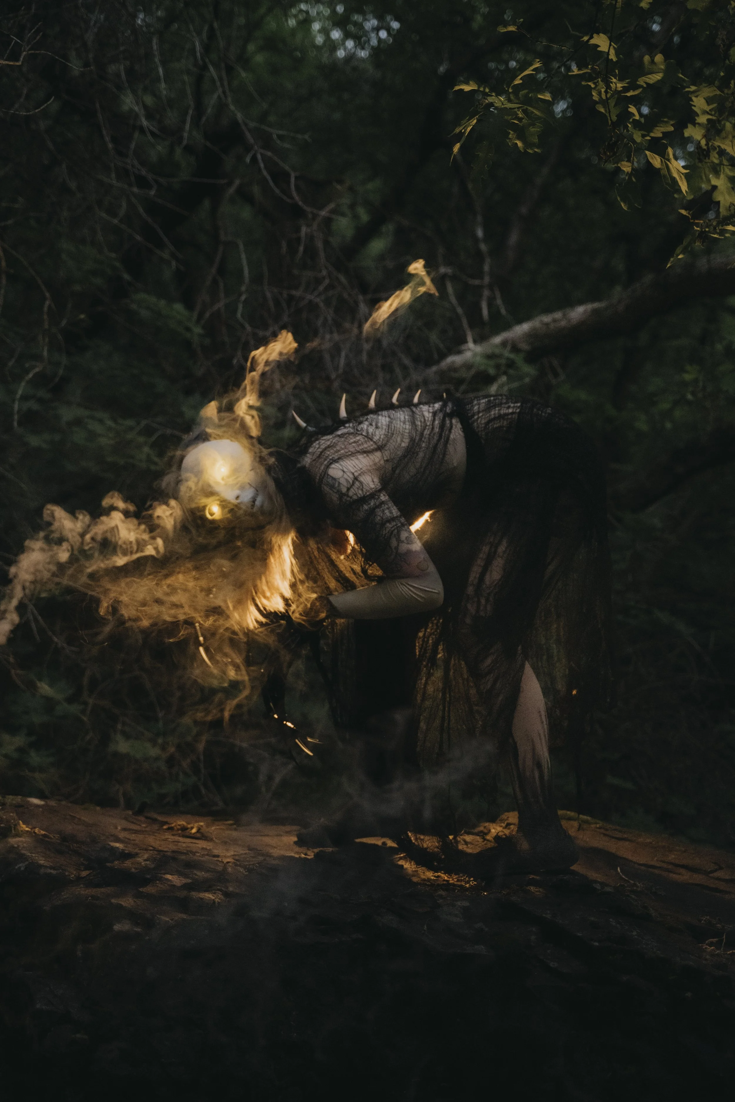 A person with long dark hair and a black mesh dress, wearing a horned mask, is kneeling and tending to a small fire at night in a wooded area. The surroundings are dark with trees and foliage.