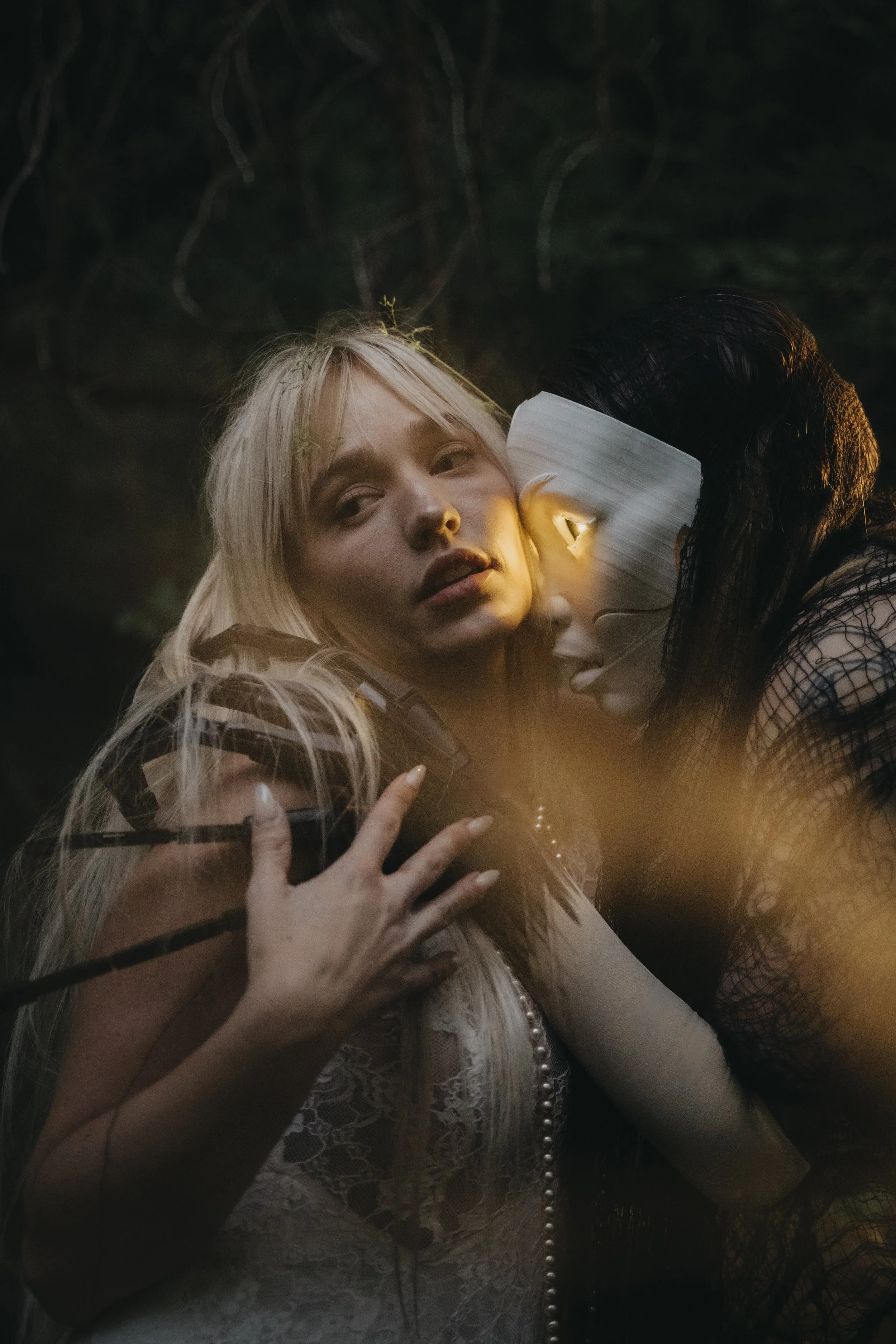 A woman with long blonde hair and a string of pearls around her neck holds a glass and gazes thoughtfully into the distance, with a dark background and faint trees visible.