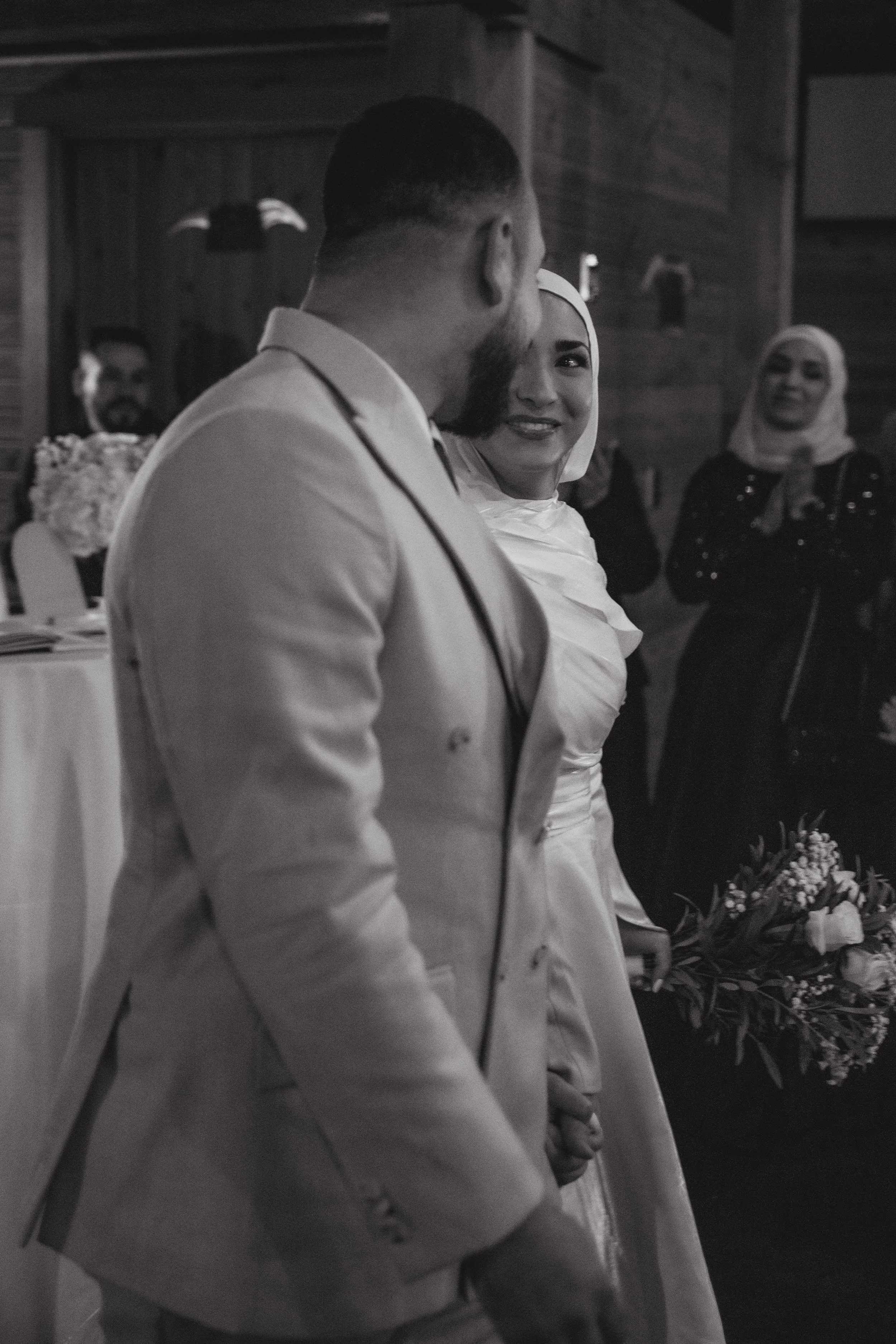 A bride and groom at their wedding, holding hands and looking at each other in a rustic indoor setting, with guests in the background.