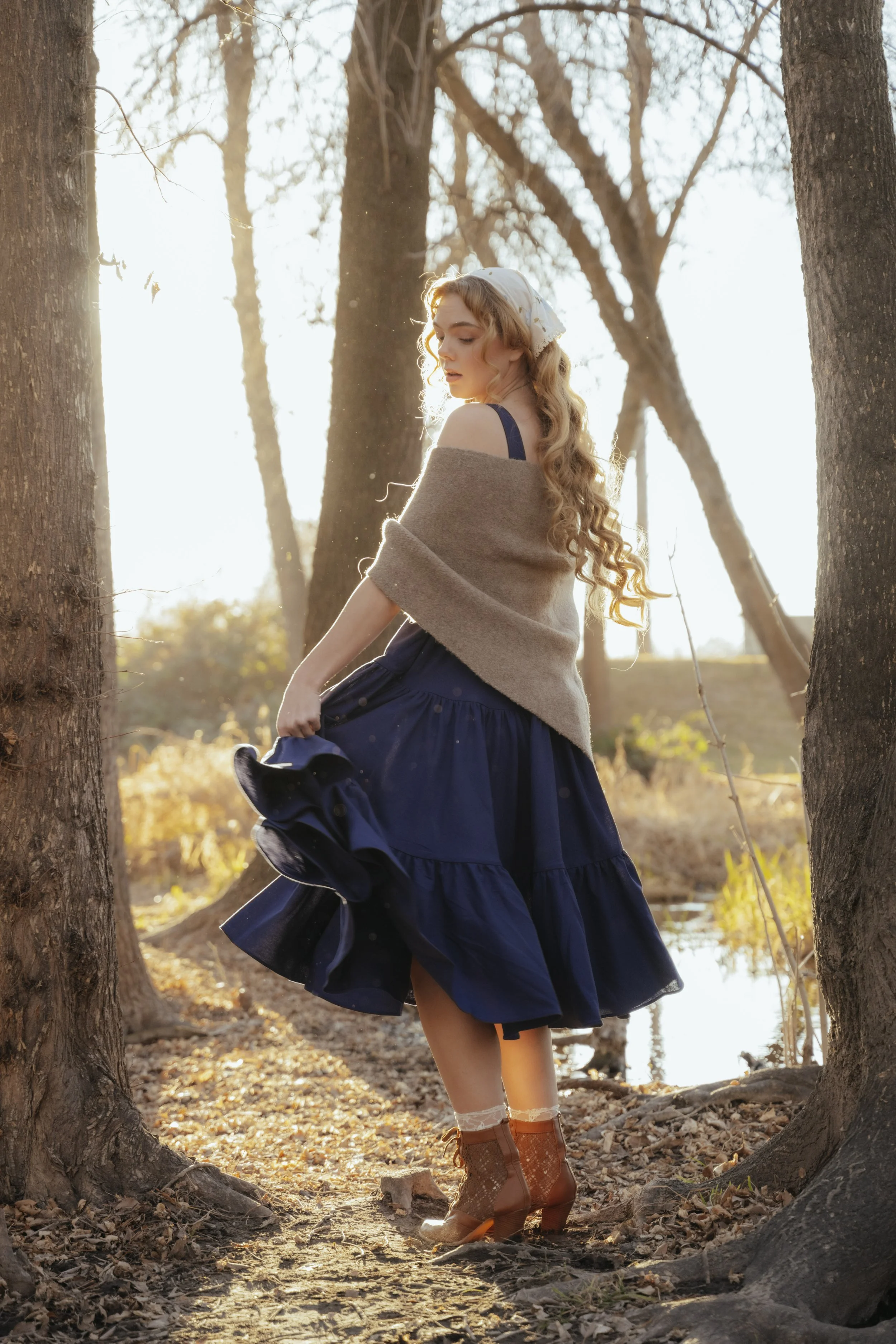 A young woman with long, curly blonde hair, wearing a dark blue dress, a beige shawl, and brown ankle boots, standing between two trees in a wooded area during sunset. She is holding the hem of her dress, lifting it slightly, as she looks downward.
