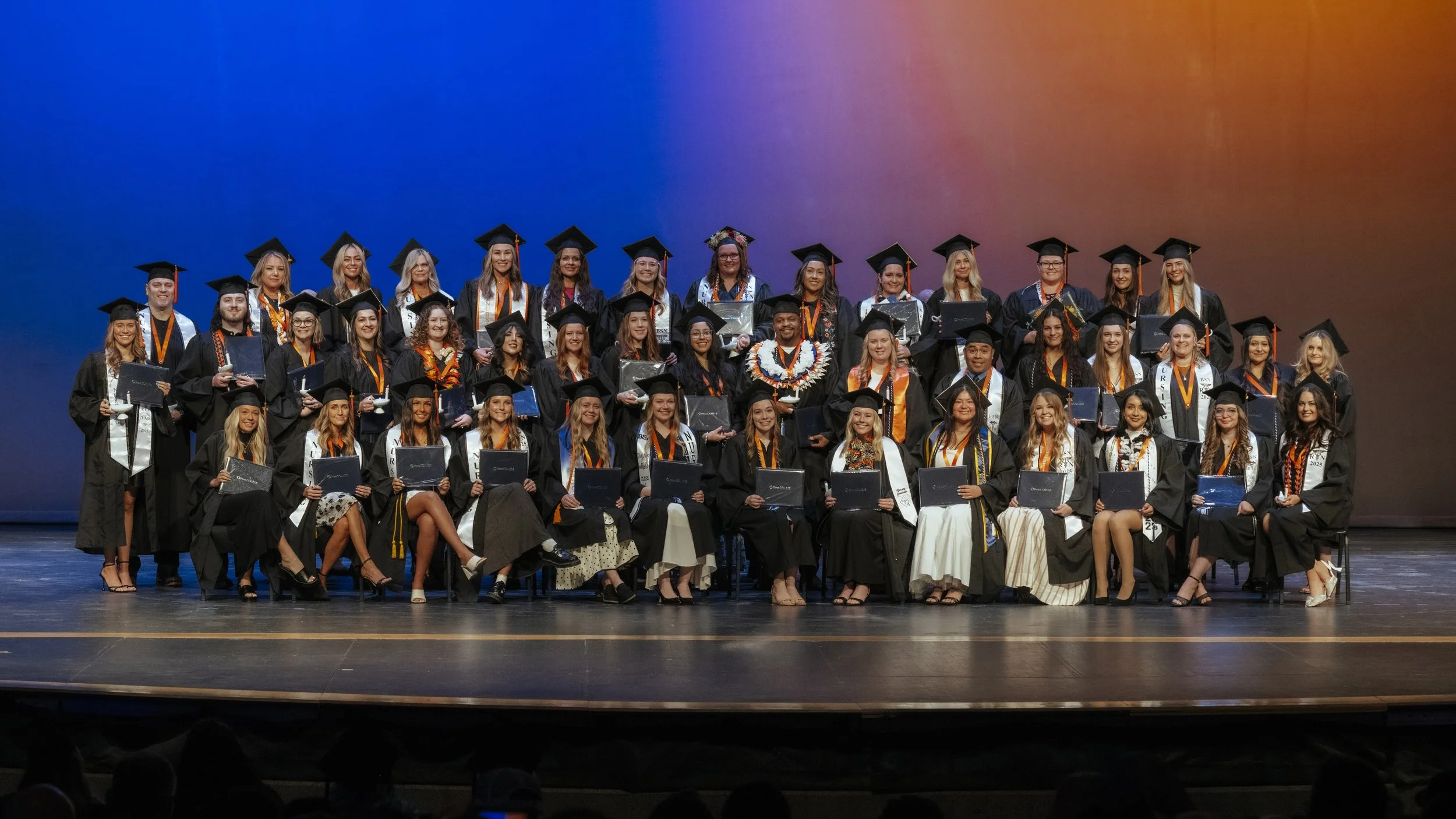 Group of students in caps and gowns celebrating graduation on stage with a colorful backdrop.