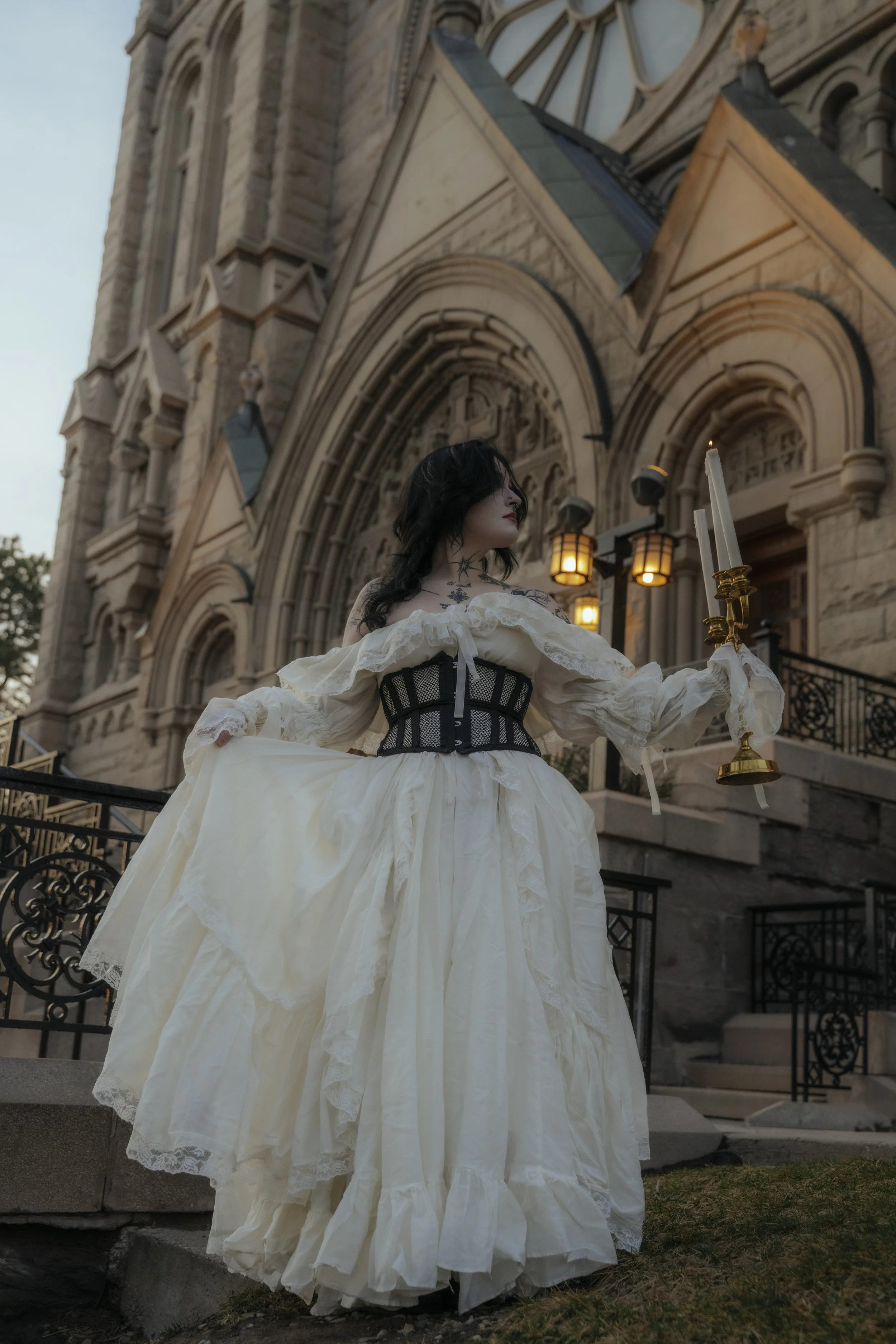A woman dressed in a Victorian-style cream gown with a black corset stands in front of an old stone church holding a gold candlestick with lit candles. The church has gothic architecture with stained glass windows and ornate stonework, illuminated by