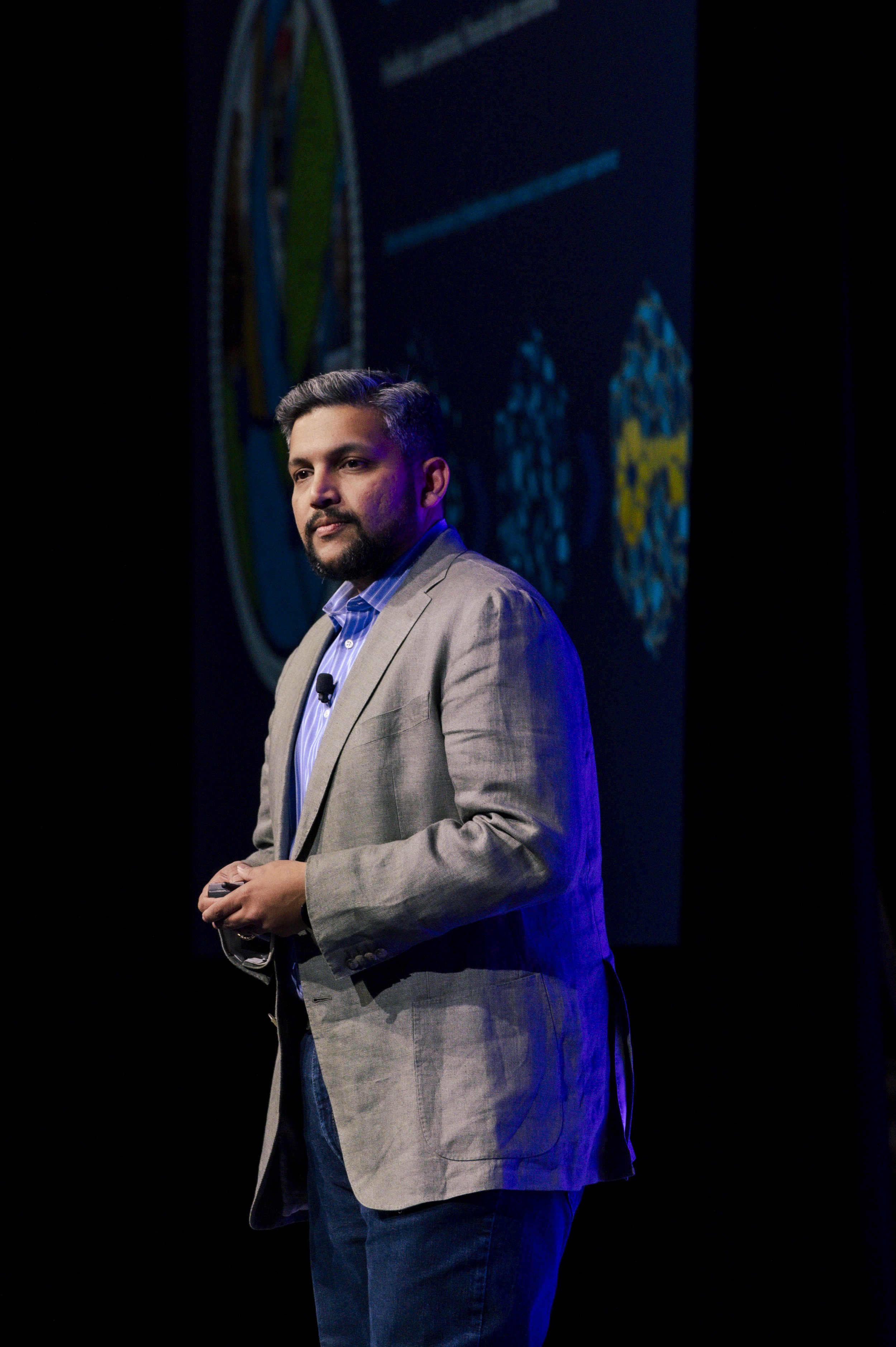 Man in light gray blazer and blue shirt giving a presentation on stage with a dark background and a large screen displaying graphics.
