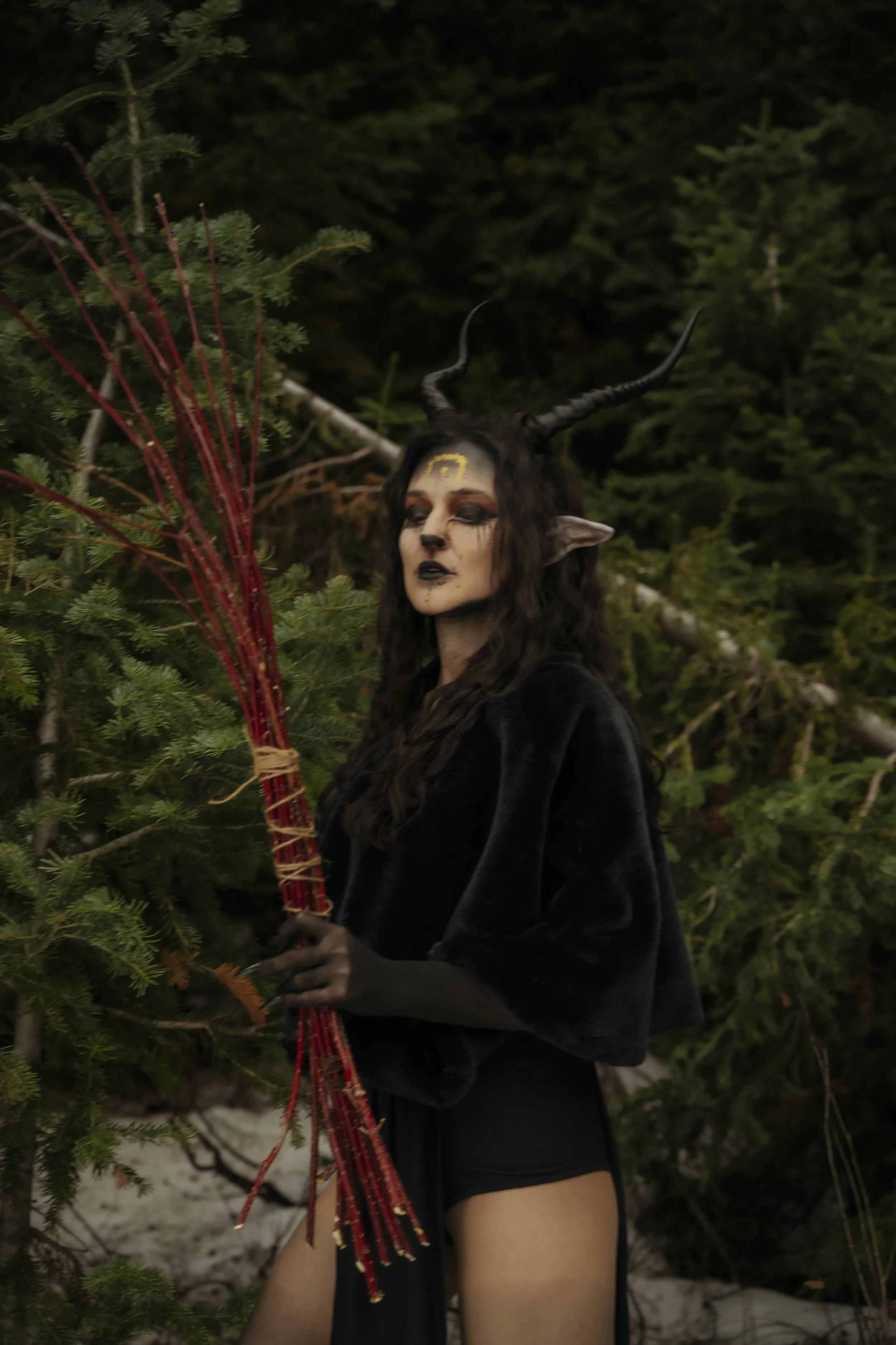 A person dressed as a mythological or fantasy character with horned headgear, dark makeup, and elf-like ears, holding a bundle of red decorative sticks, standing in a forested area.
