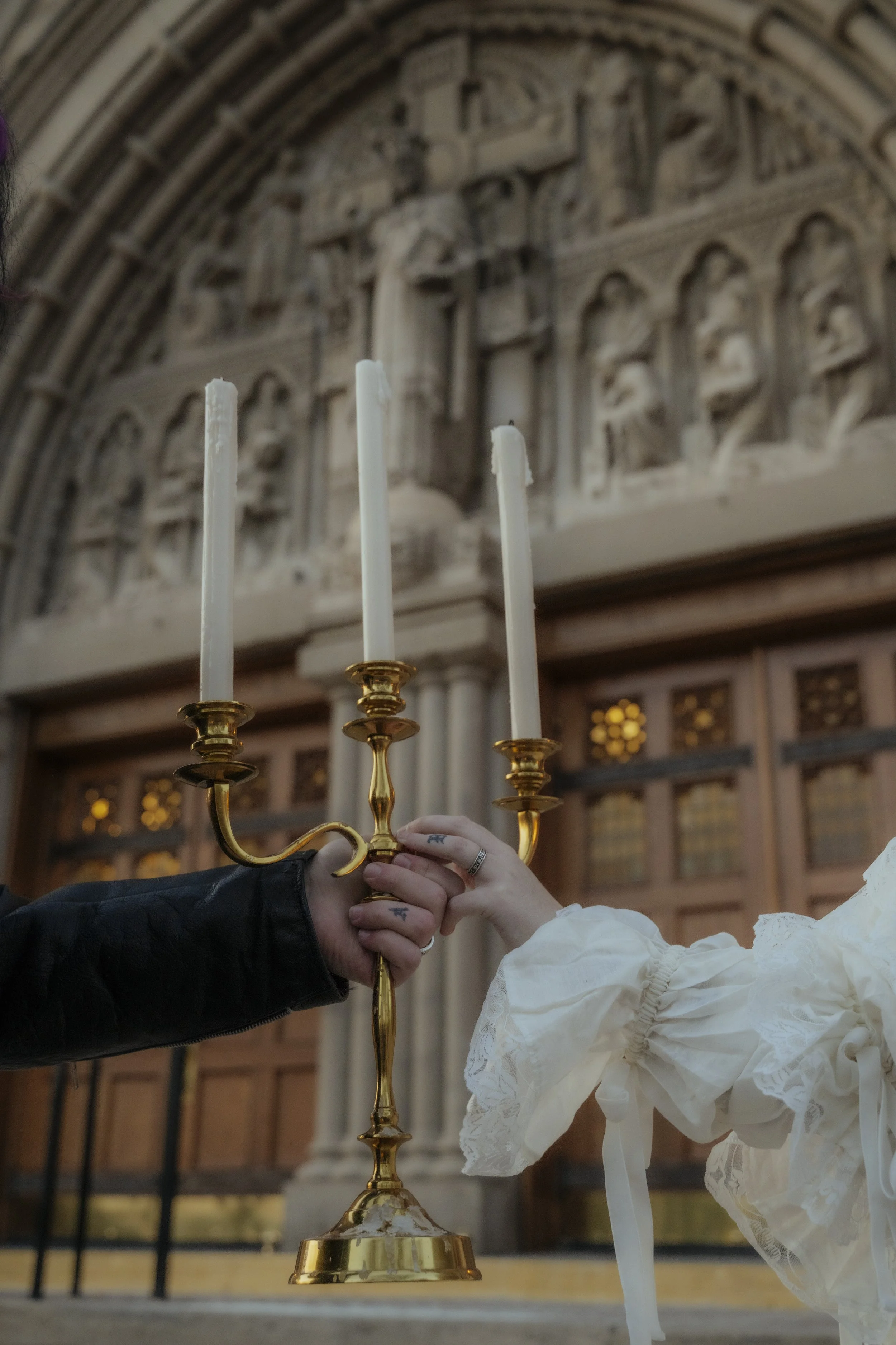 Two hands, one with tattoos and rings, holding a gold candelabrum with three white candles, inside a church with ornate woodwork and stone carvings in the background.