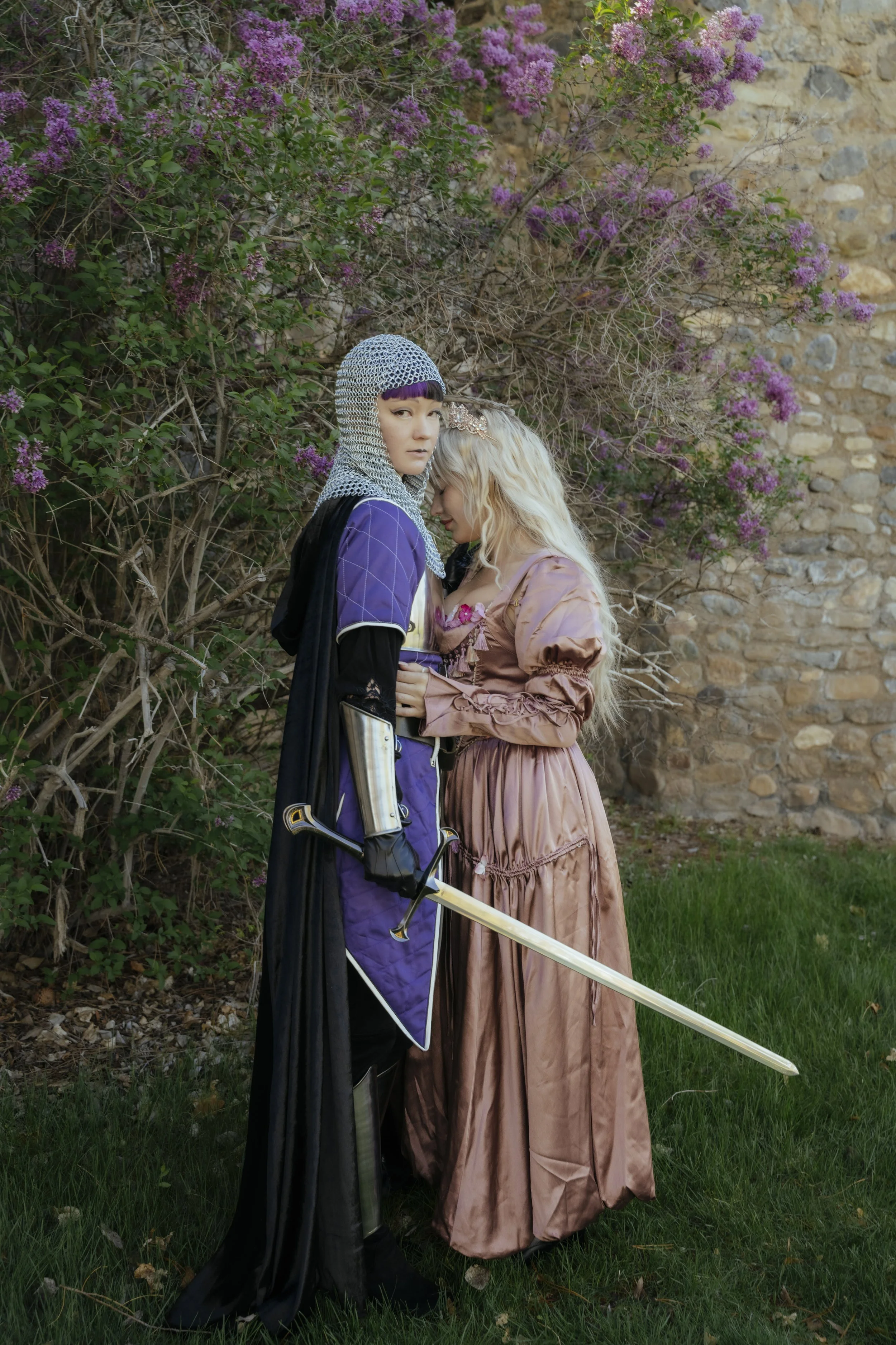 Two women dressed as fantasy characters stand close together outdoors near purple flowering bushes and a stone wall. One is wearing medieval armor and a chainmail hood, holding a sword, while the other is in a long, silky, pinkish-brown gown with blo