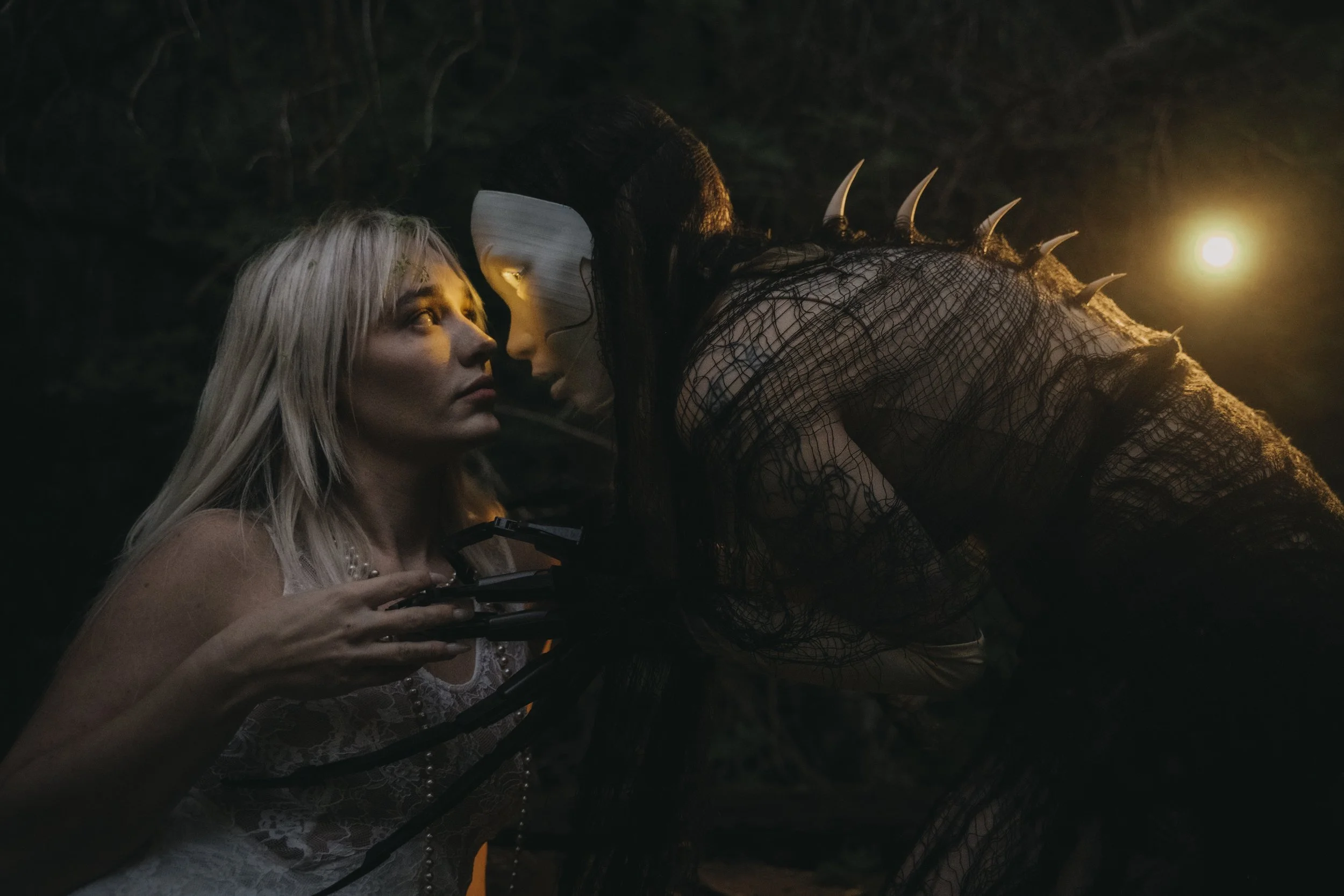 Two women, one with blond hair and one dressed as a monster with spikes on its back, are looking at a drone controller in their hands in a dark outdoor setting illuminated by a nearby light.