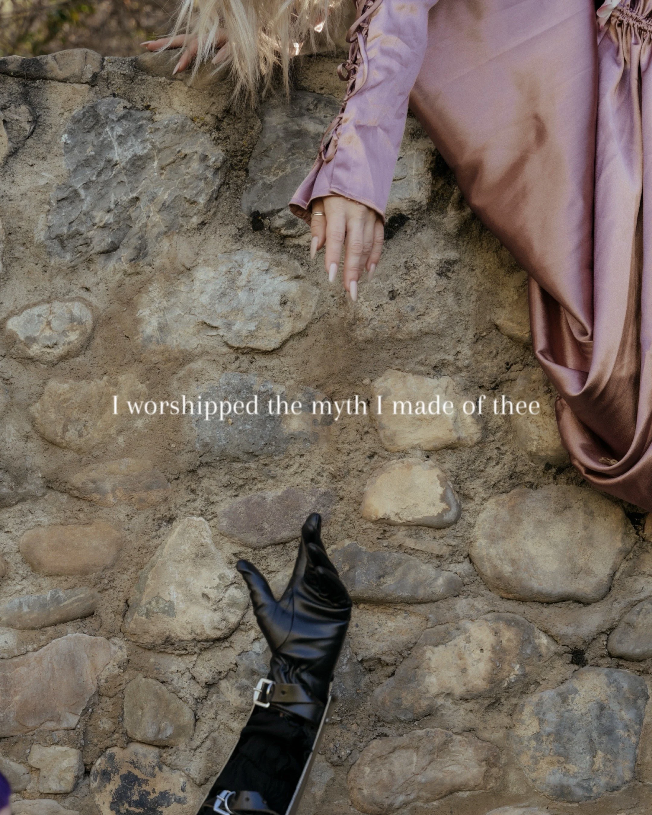 Close-up of two hands, one with a ring, reaching towards each other over a stone wall. One hand is bare, the other is covered by a black leather glove. The background shows a stone wall with a quote overlayed that reads 'I worshipped the myth I made 