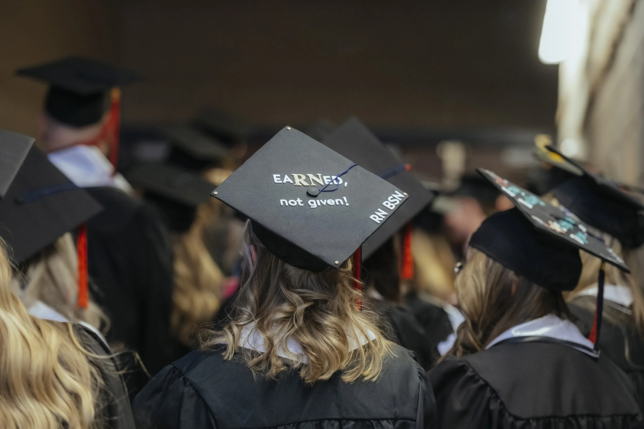 Group of people wearing black graduation caps and gowns, seated during a graduation ceremony, with several caps visible, one of which has words partially visible reading "EARNED, not given!"