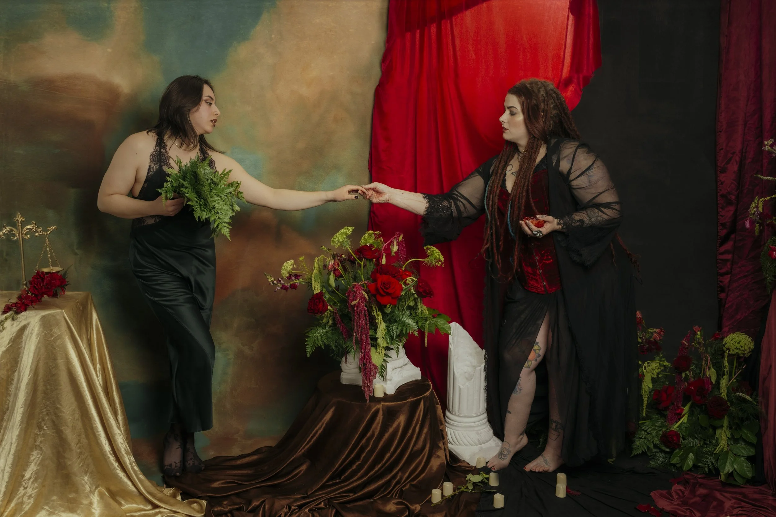 Two women in gothic attire engaging in a symbolic ritual involving a black and red color scheme, with one woman passing a red object to the other amid floral arrangements, candles, and velvet drapery.