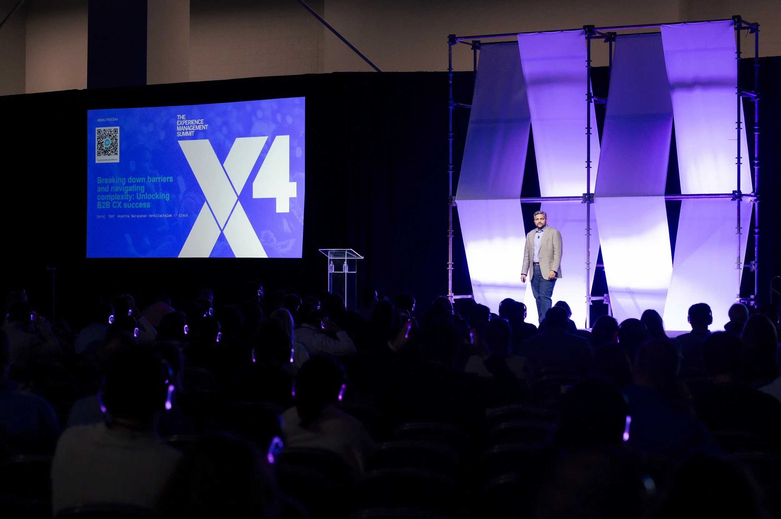 A speaker stands on stage at the Experience Management Summit, delivering a presentation to an audience wearing headphones, with a large screen displaying the Summit logo and presentation title, lit with purple and blue stage lighting.