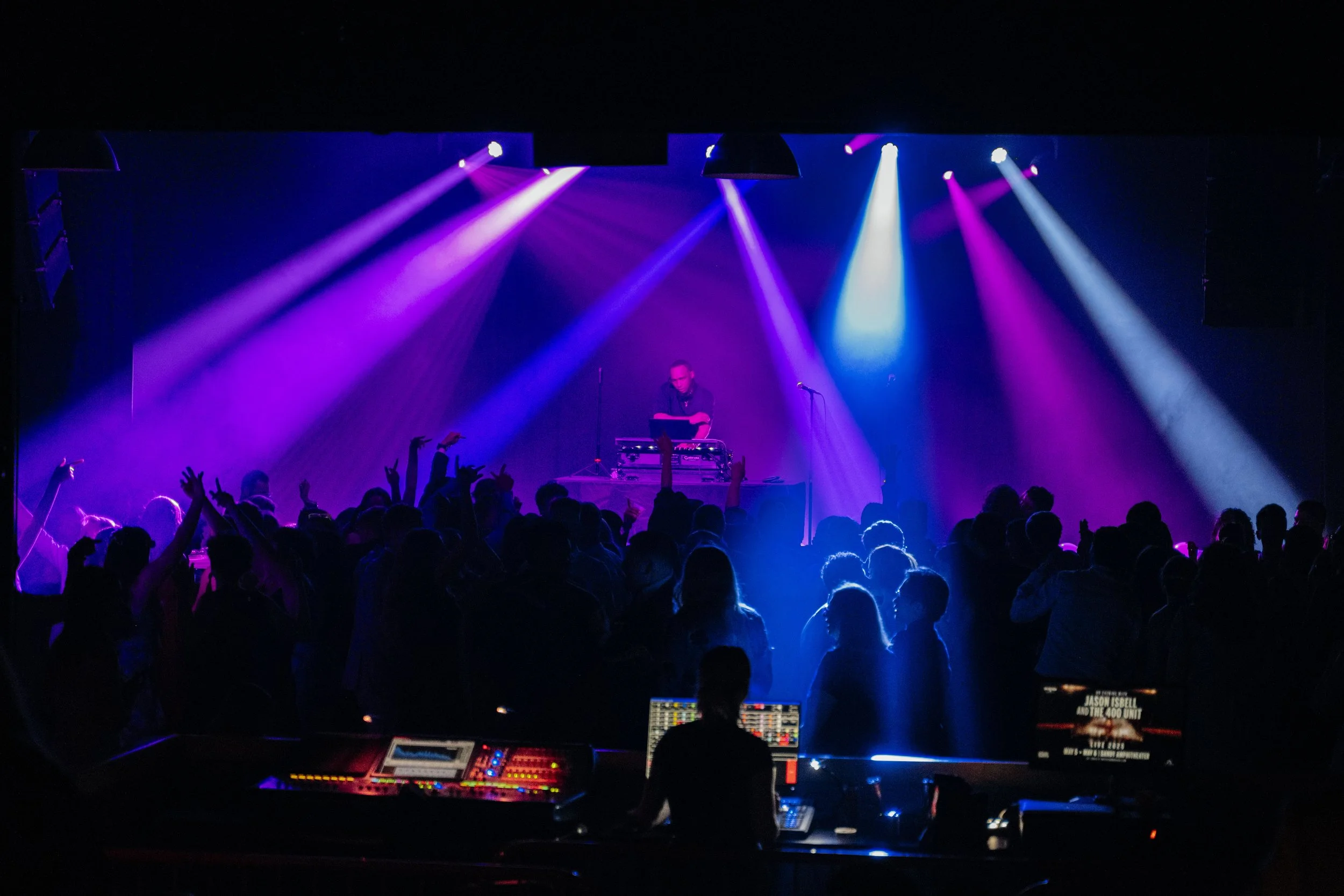 A DJ performing on stage with colorful purple and blue stage lights, crowd dancing, and audio equipment in the foreground.