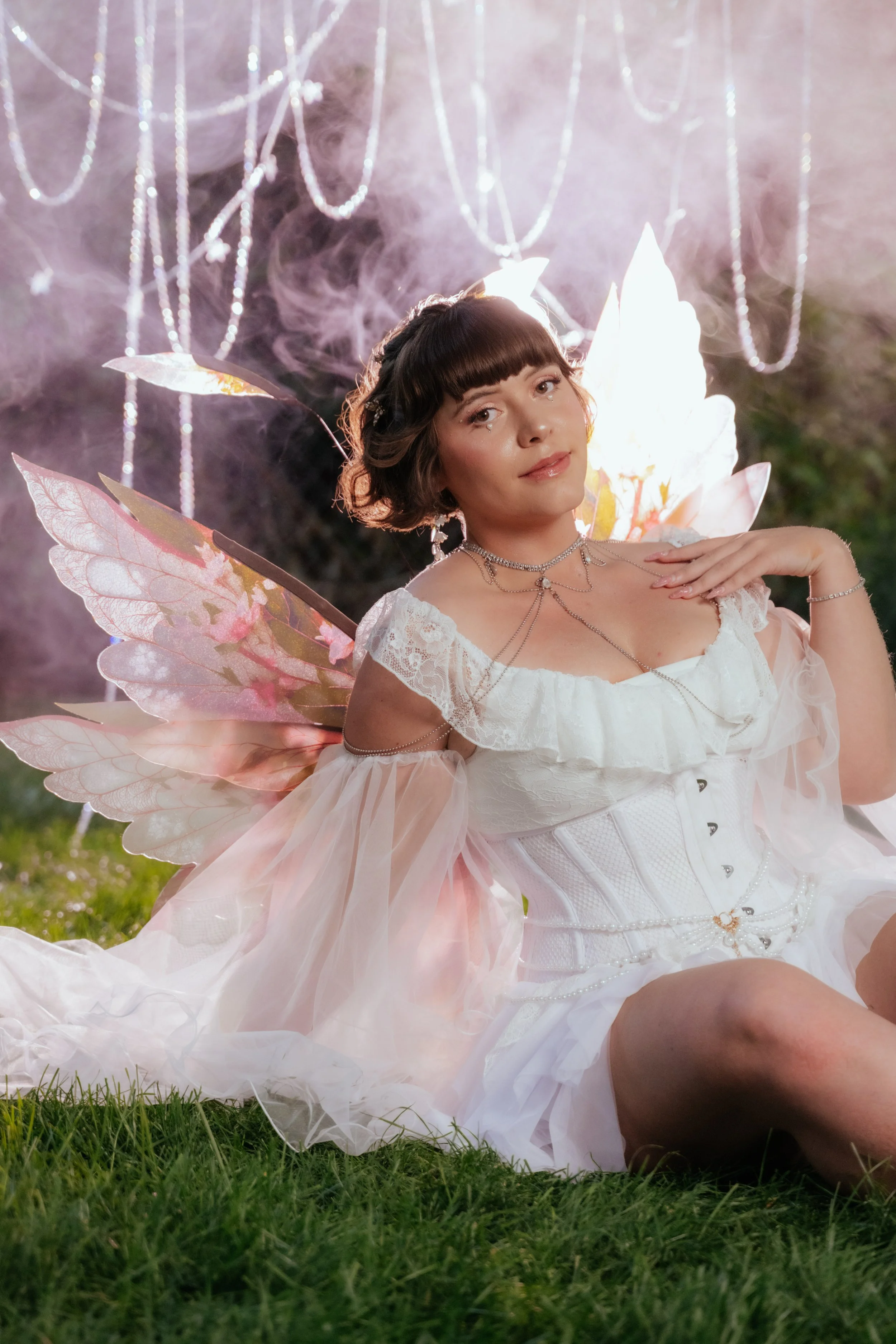 A woman dressed as a fairy with iridescent wings, wearing a white dress and jewelry, sitting on grass at night with mist and hanging lights in the background.
