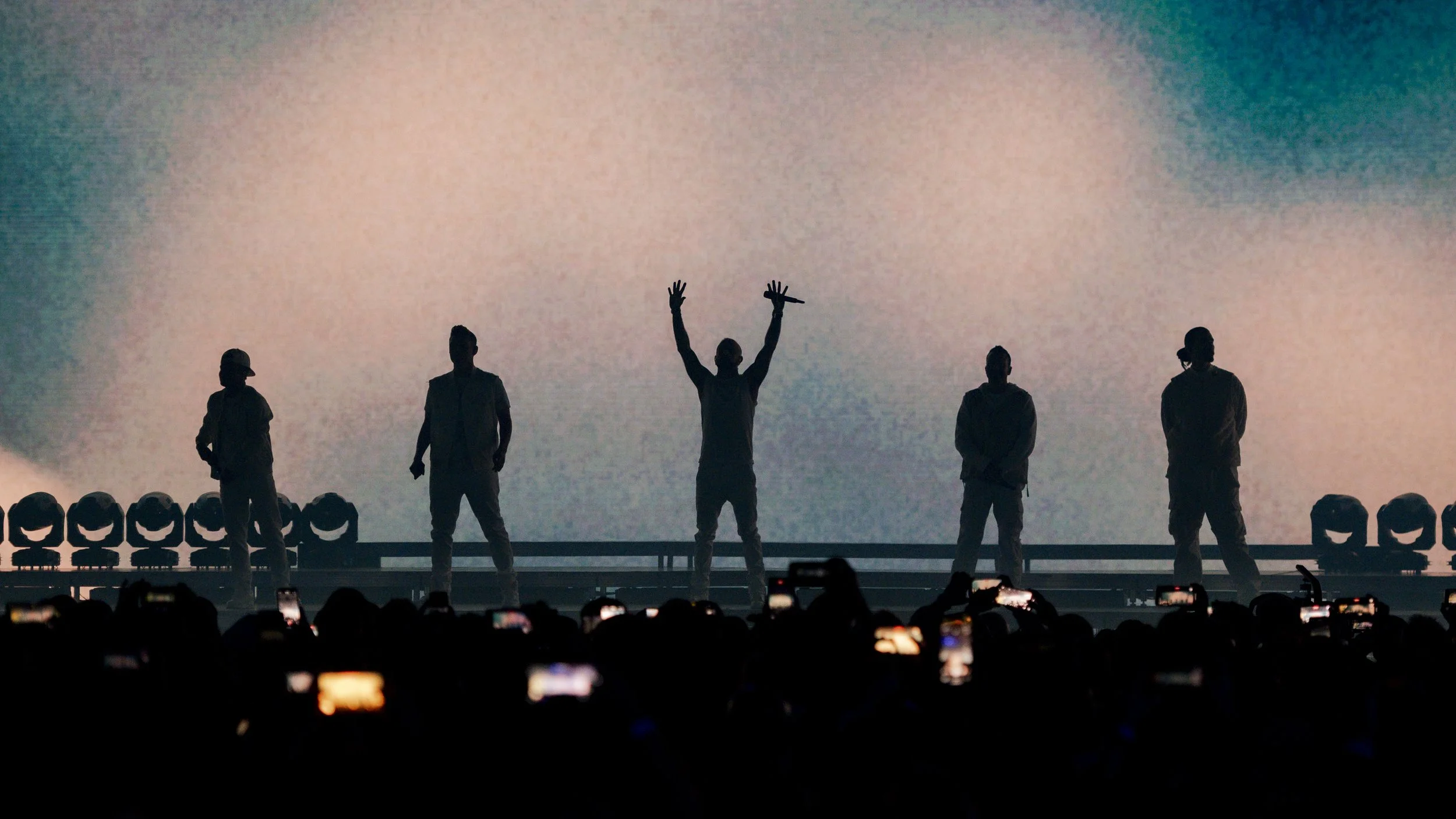 Silhouettes of five performers on stage, with one holding a microphone, against a large, colorful screen backdrop. Audience members are visible in the foreground, capturing the moment with phones.