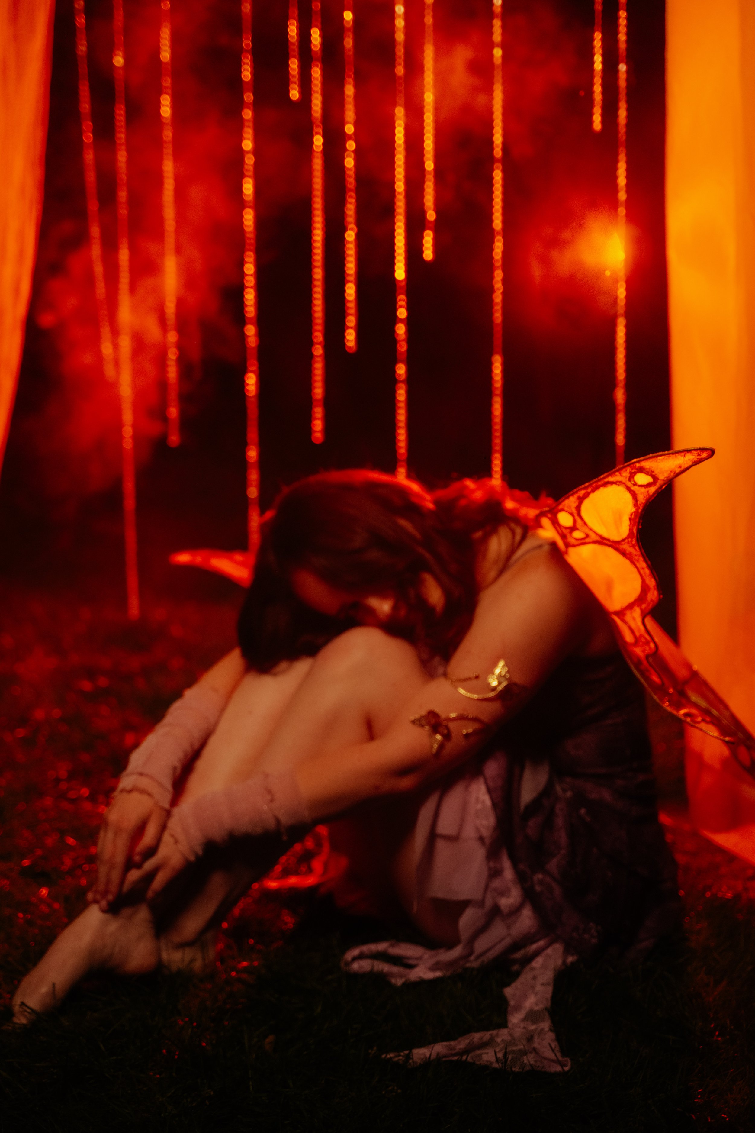 A person dressed as a fairy with orange wings hugging another person, with a dark background illuminated by red-orange vertical light streaks and curtains.