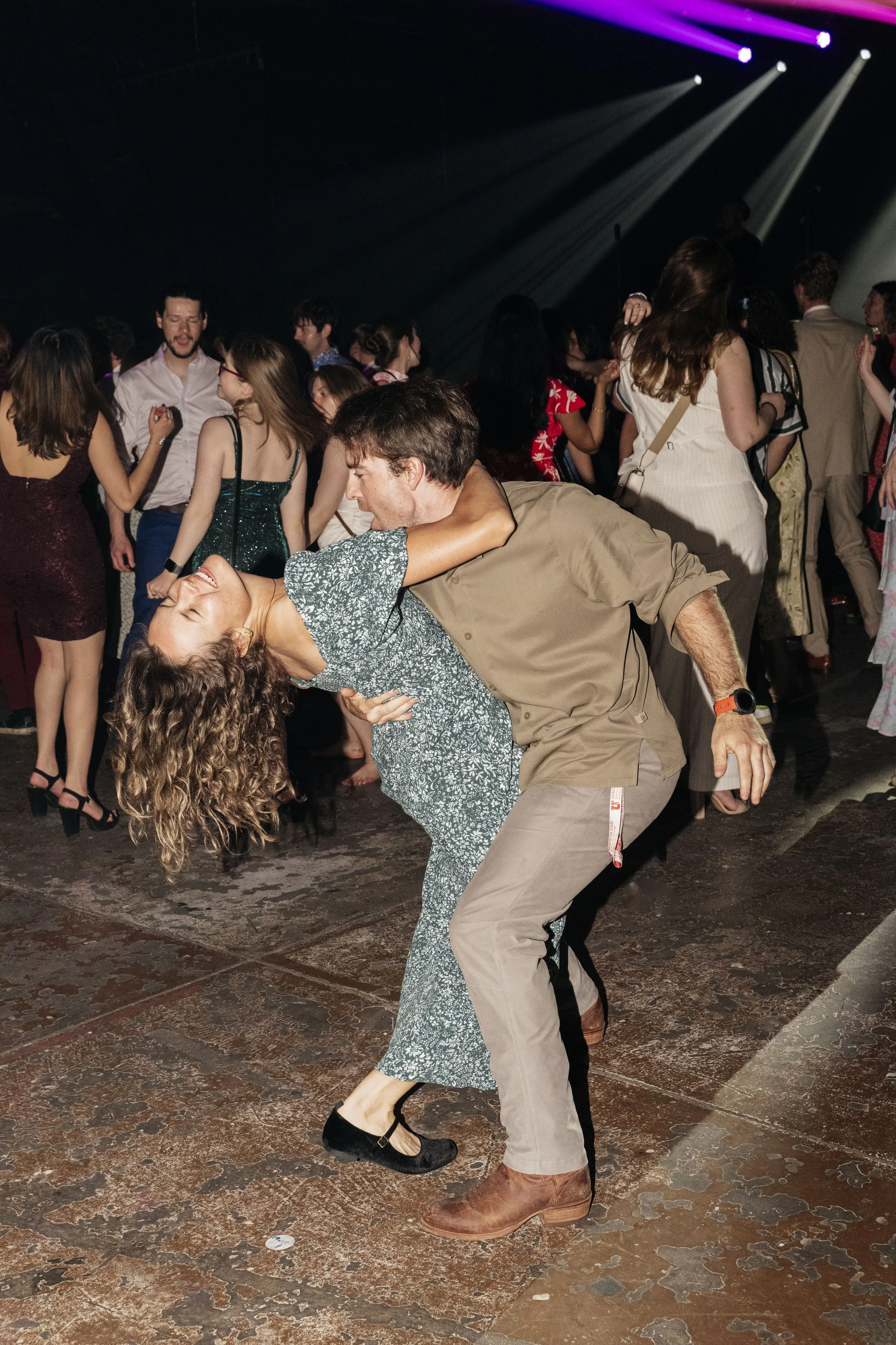 A man and woman dancing closely on a crowded dance floor, with other people dancing in the background, under colorful stage lighting.