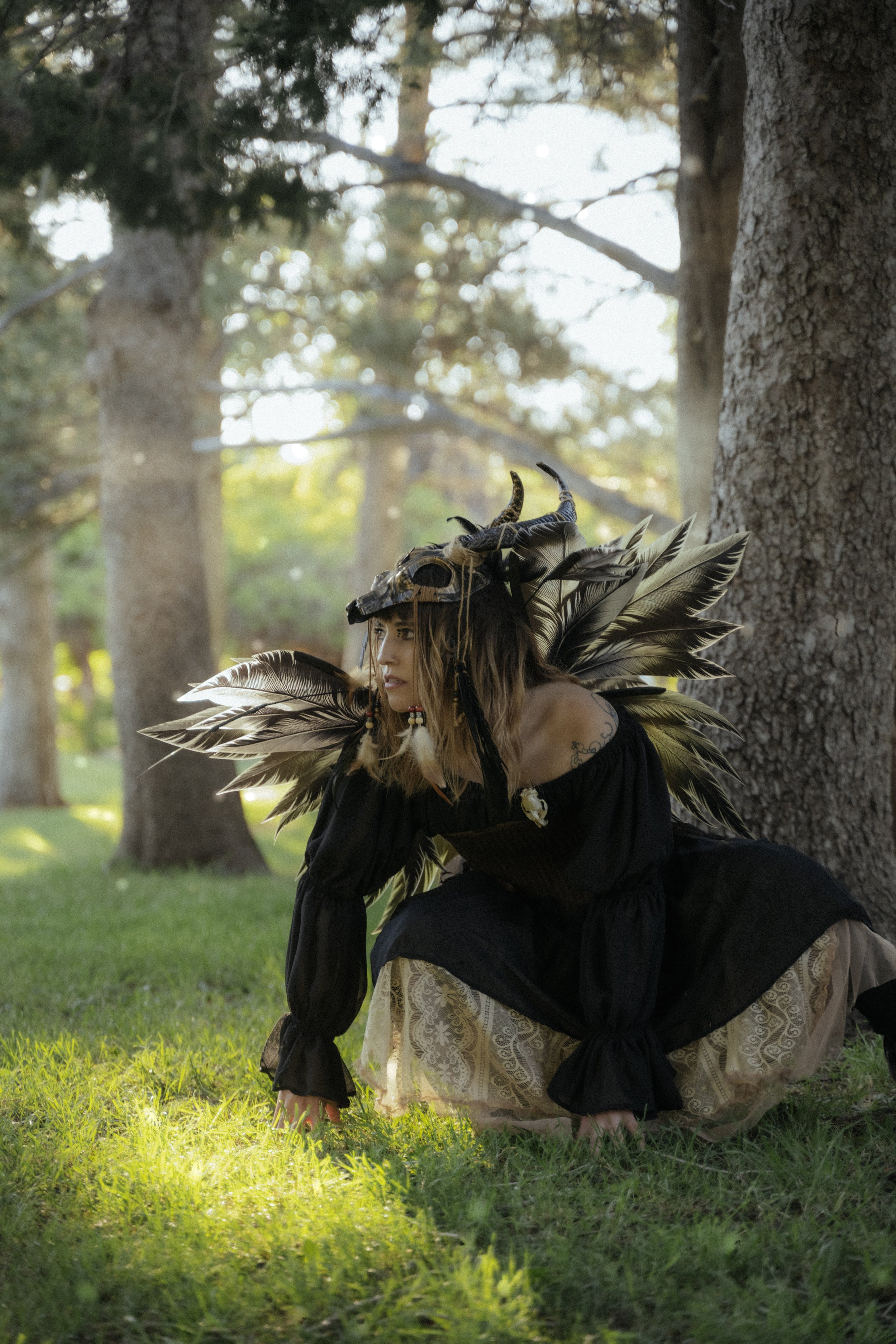 A woman dressed in tribal or fantasy costume crouches on the grass next to a large tree, wearing an elaborate feathered headdress and attire, with a forest background.