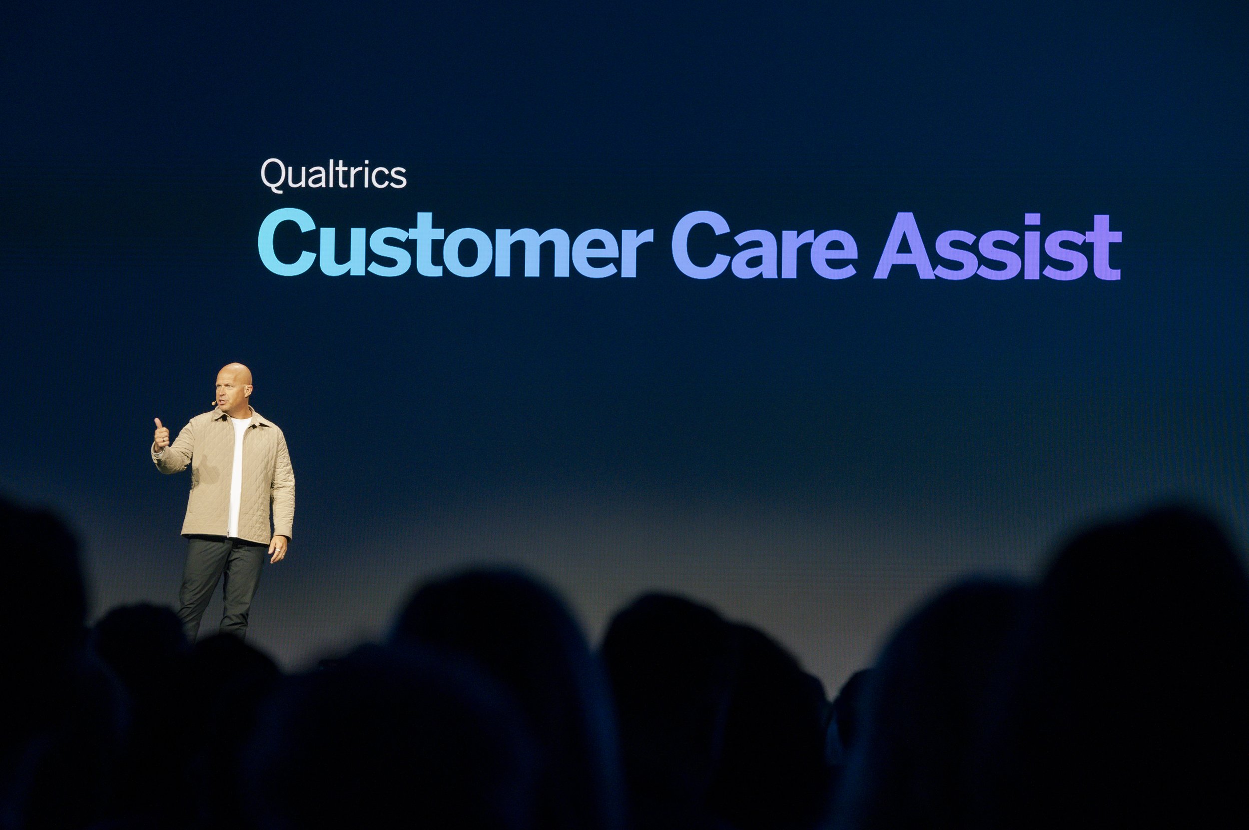 A man standing on stage giving a presentation with a large screen behind him displaying the text 'Qualtrics Customer Care Assist.'