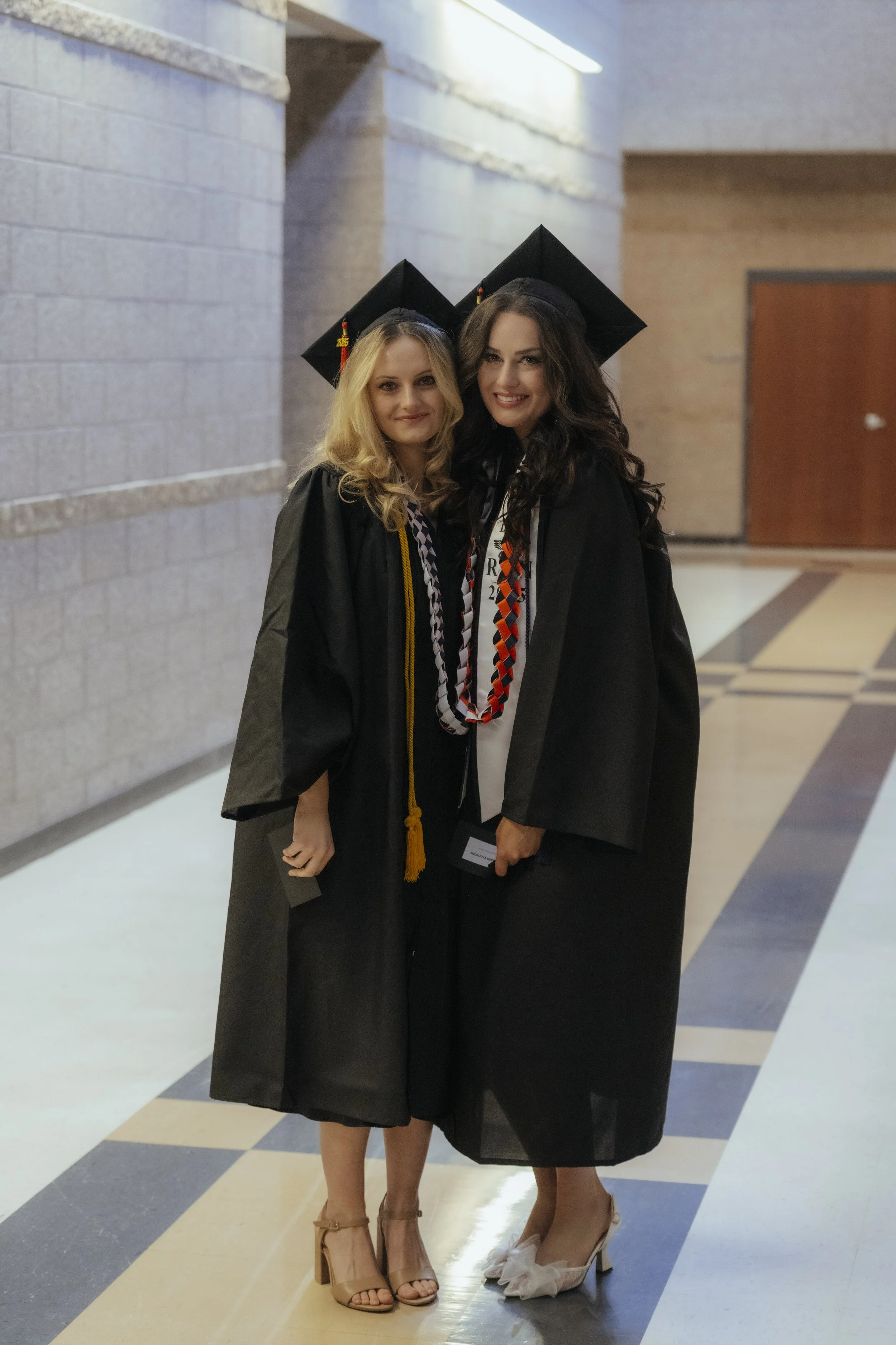 Two young women in graduation caps and gowns standing indoors, celebrating graduation, smiling at the camera.