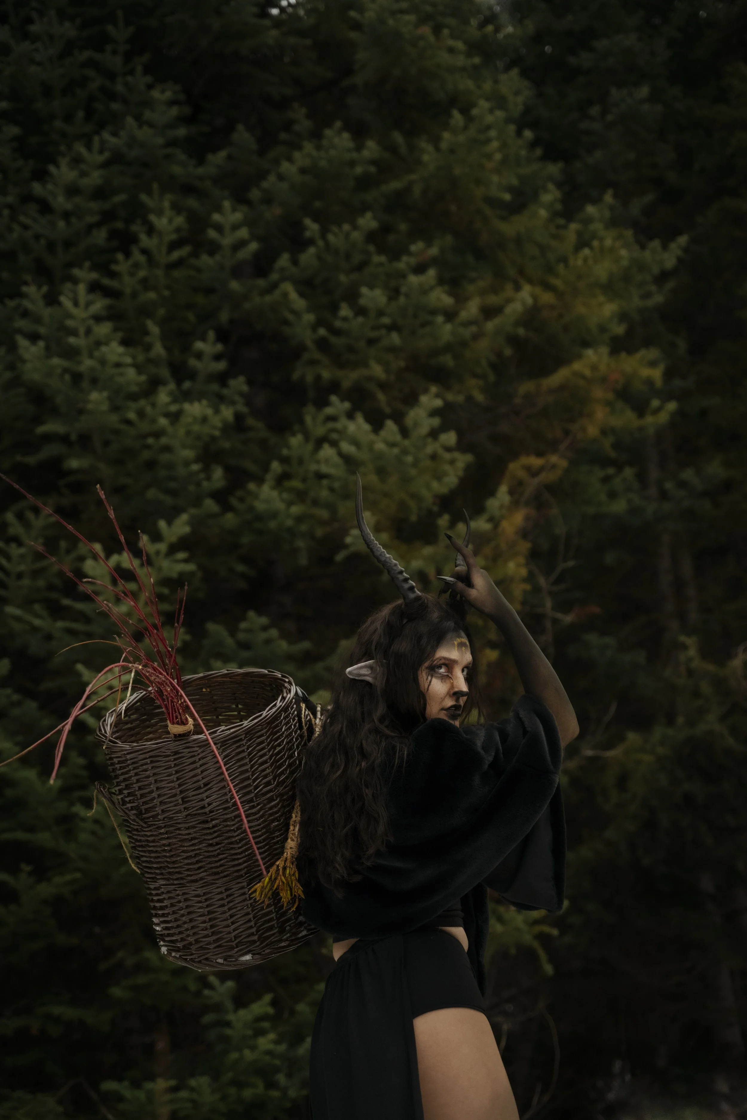 A woman with long dark hair and elaborate makeup, wearing a black outfit, stands outdoors in front of a dense forest. She has horns on her head, pointed elf ears, and is holding one horn with her right hand. She carries a woven basket on her back fil