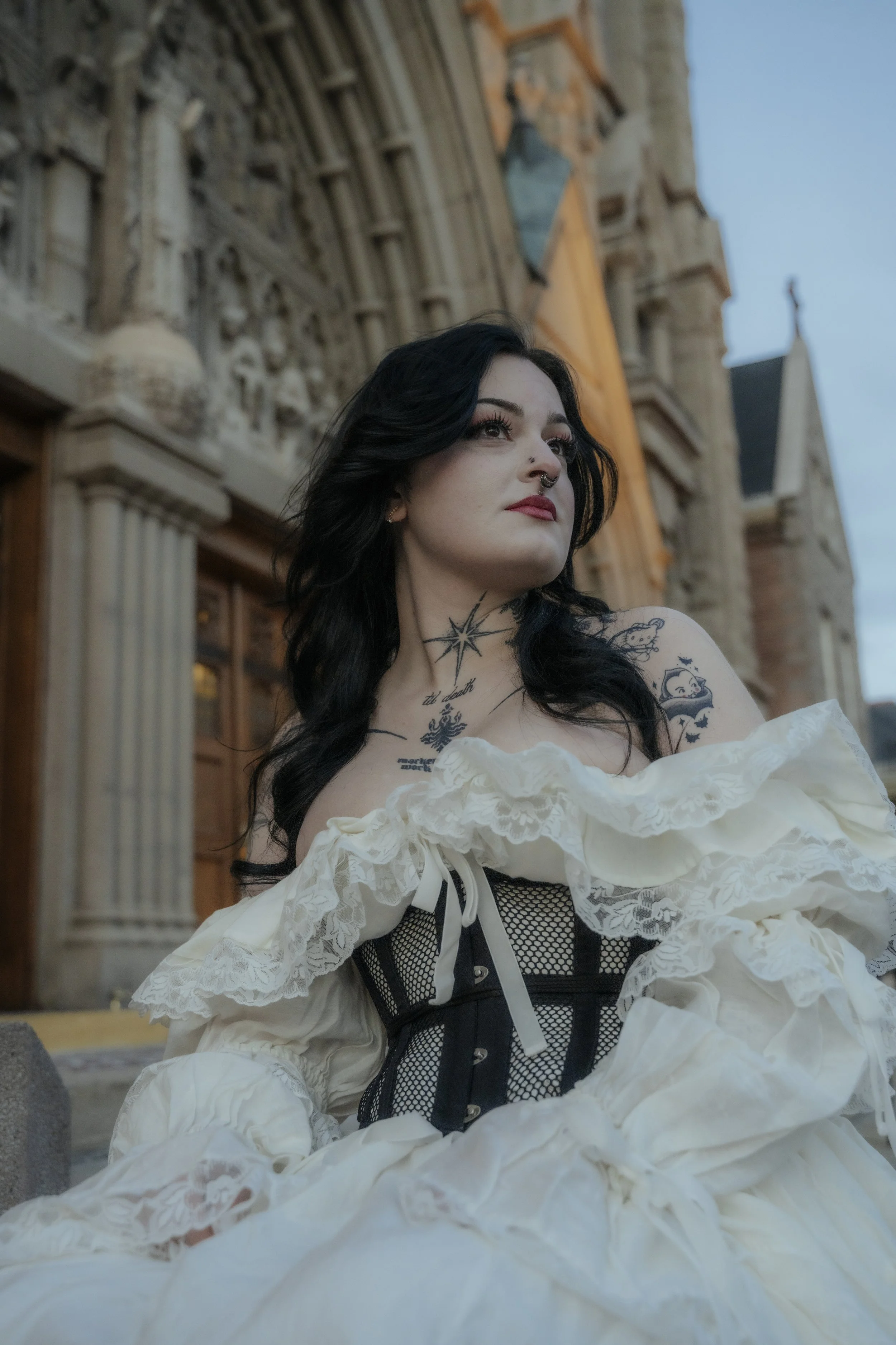 A woman with dark hair and tattoos sitting in front of a historic stone building, wearing a vintage white dress with black corset details.