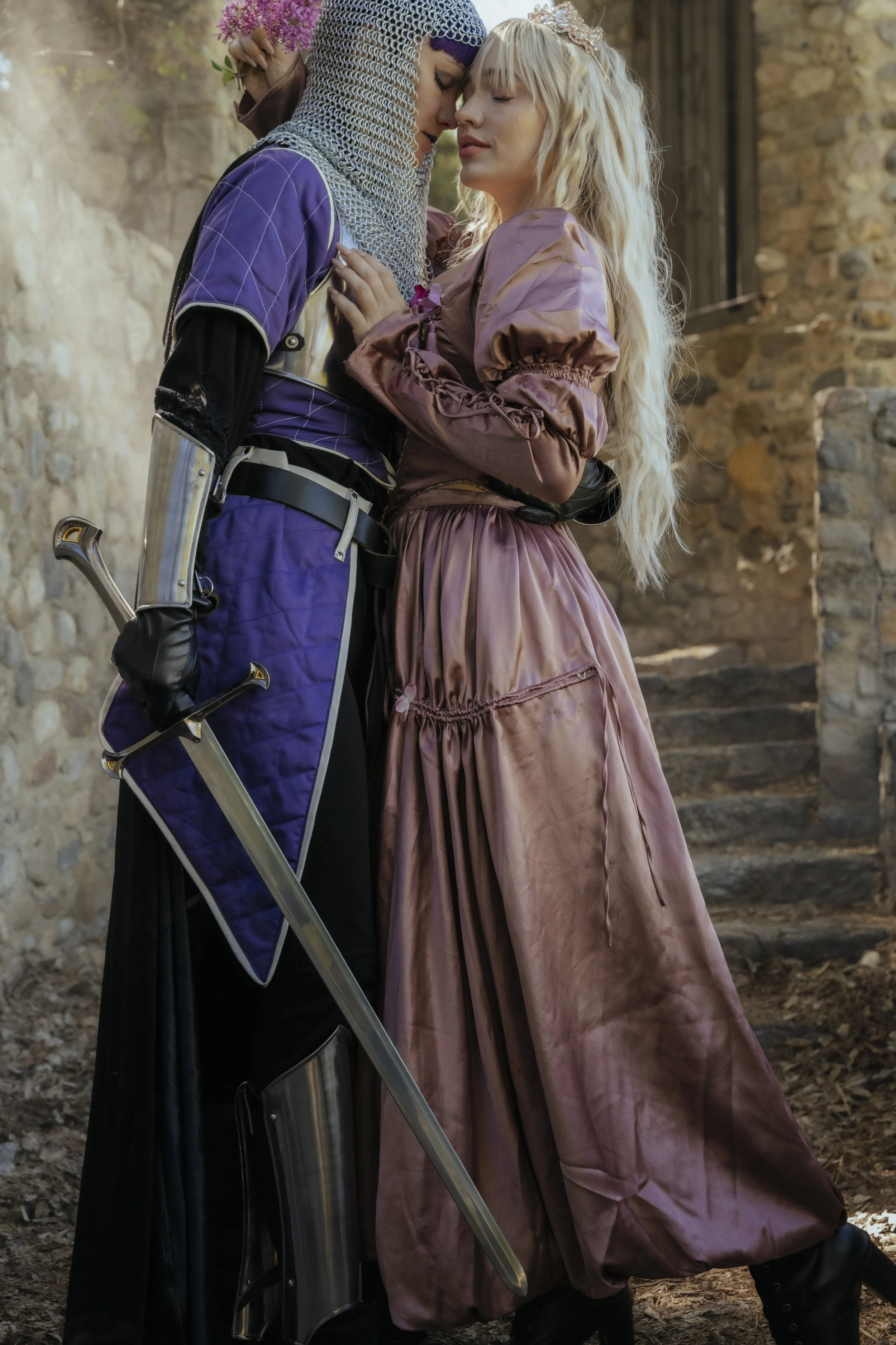 Two women in medieval costumes embrace each other outdoors near stone steps and a brick wall.