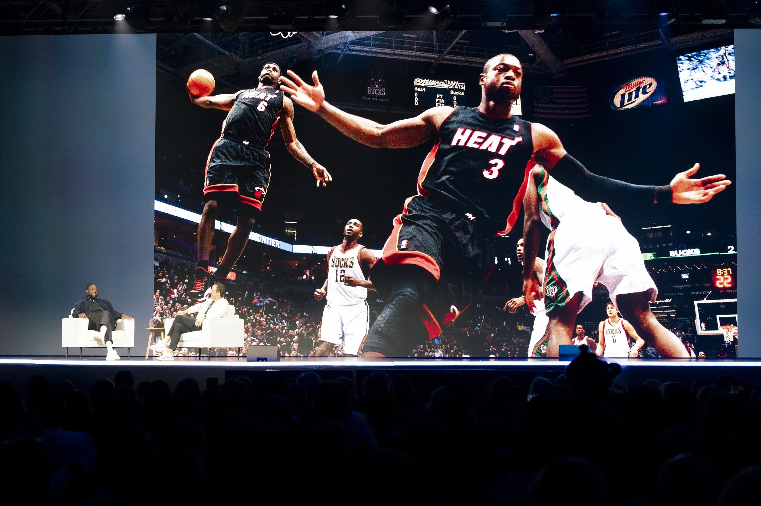 Large screen showing a basketball game featuring Miami Heat players in black jerseys with red accents on a basketball court with an audience in the dark; speakers and panelists are seated on stage in front of the screen.