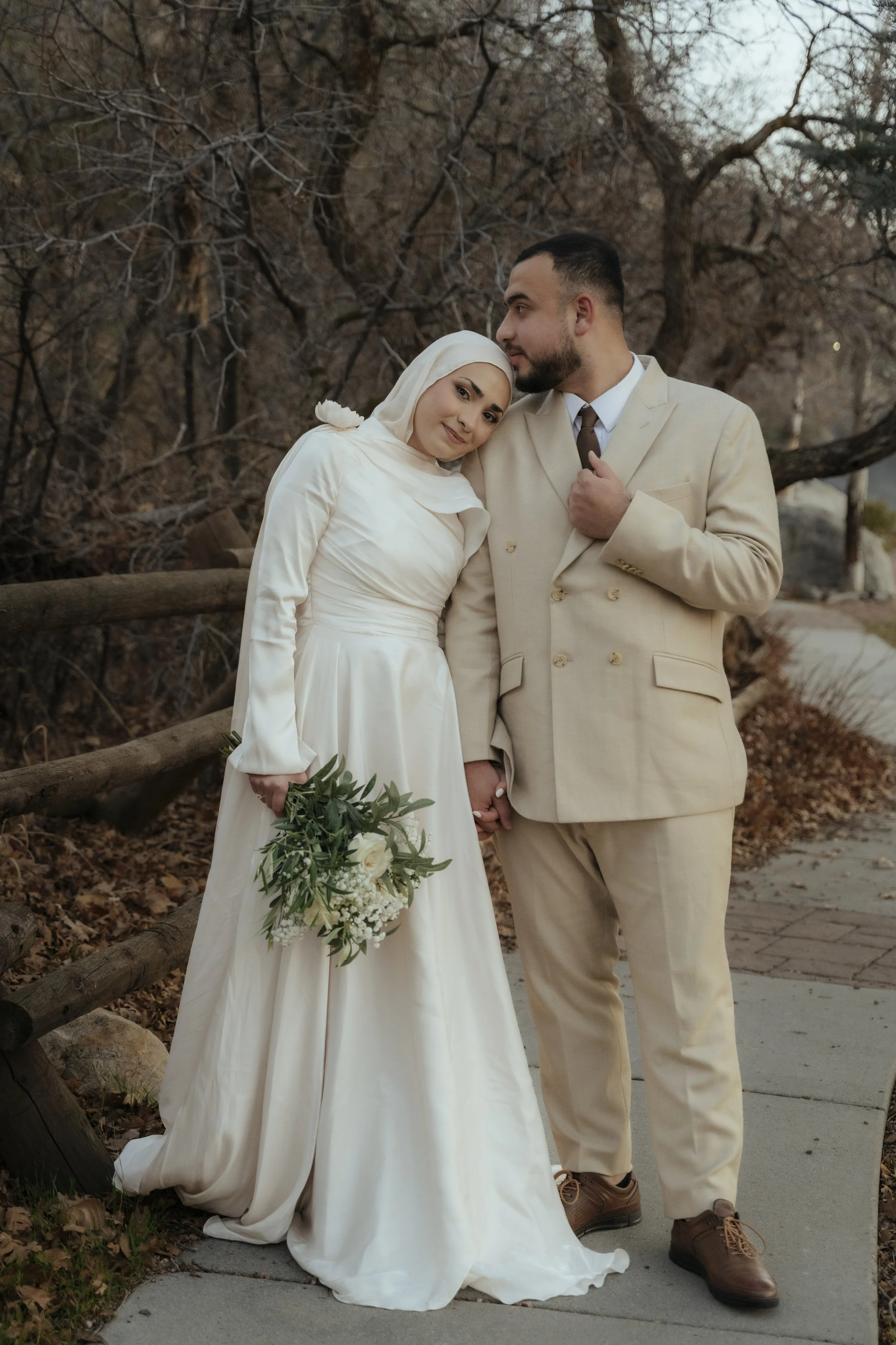 A bride and groom holding hands outdoors during fall, with bare trees and a wooden fence in the background. The bride wears a long white dress and hijab, holding a bouquet, and the groom wears a cream-colored suit.