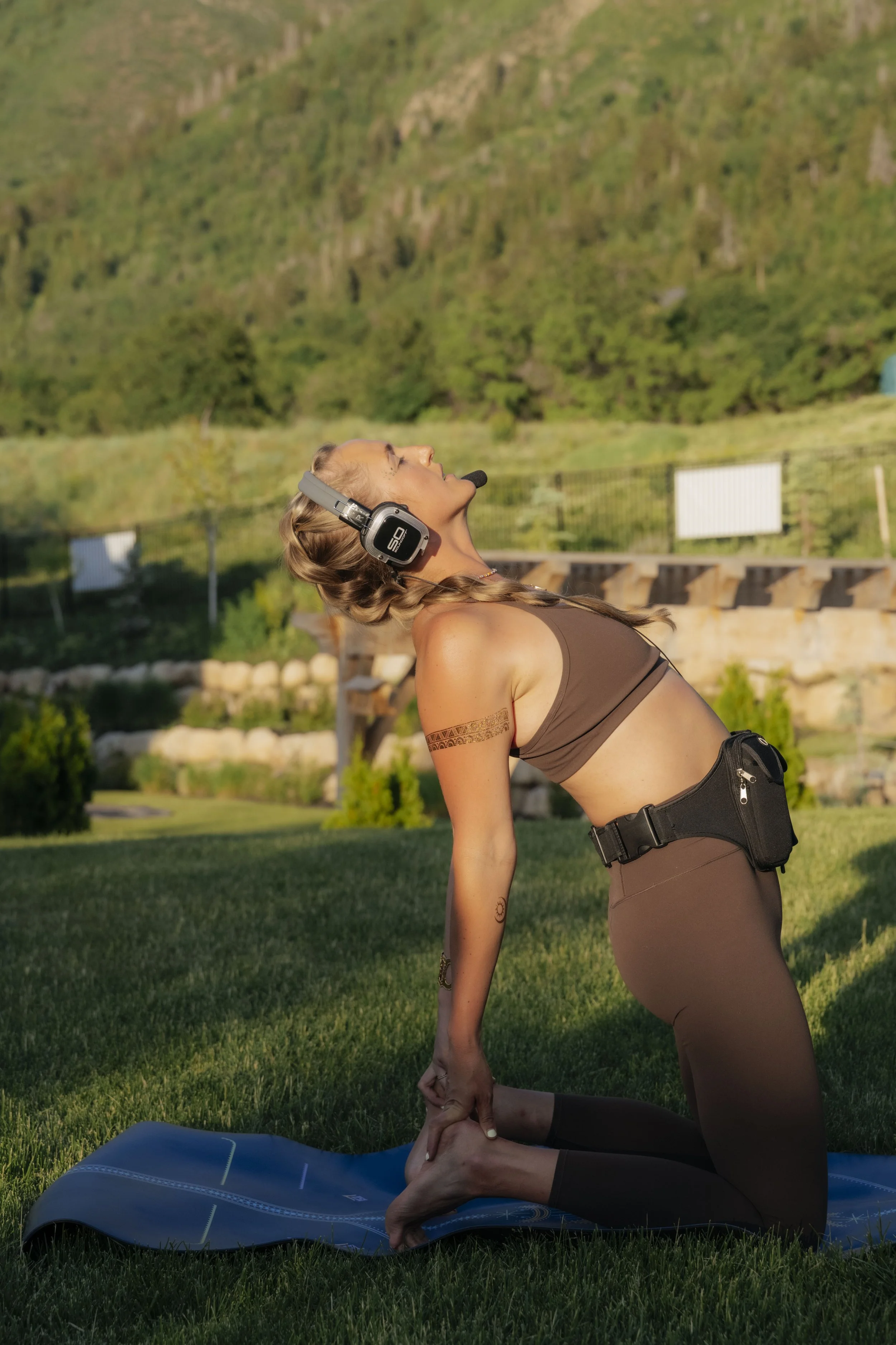 A woman practicing yoga outdoors on a grassy field, performing a lunge pose while wearing a headset, brown sports bra, and brown leggings, with a scenic green mountain background.