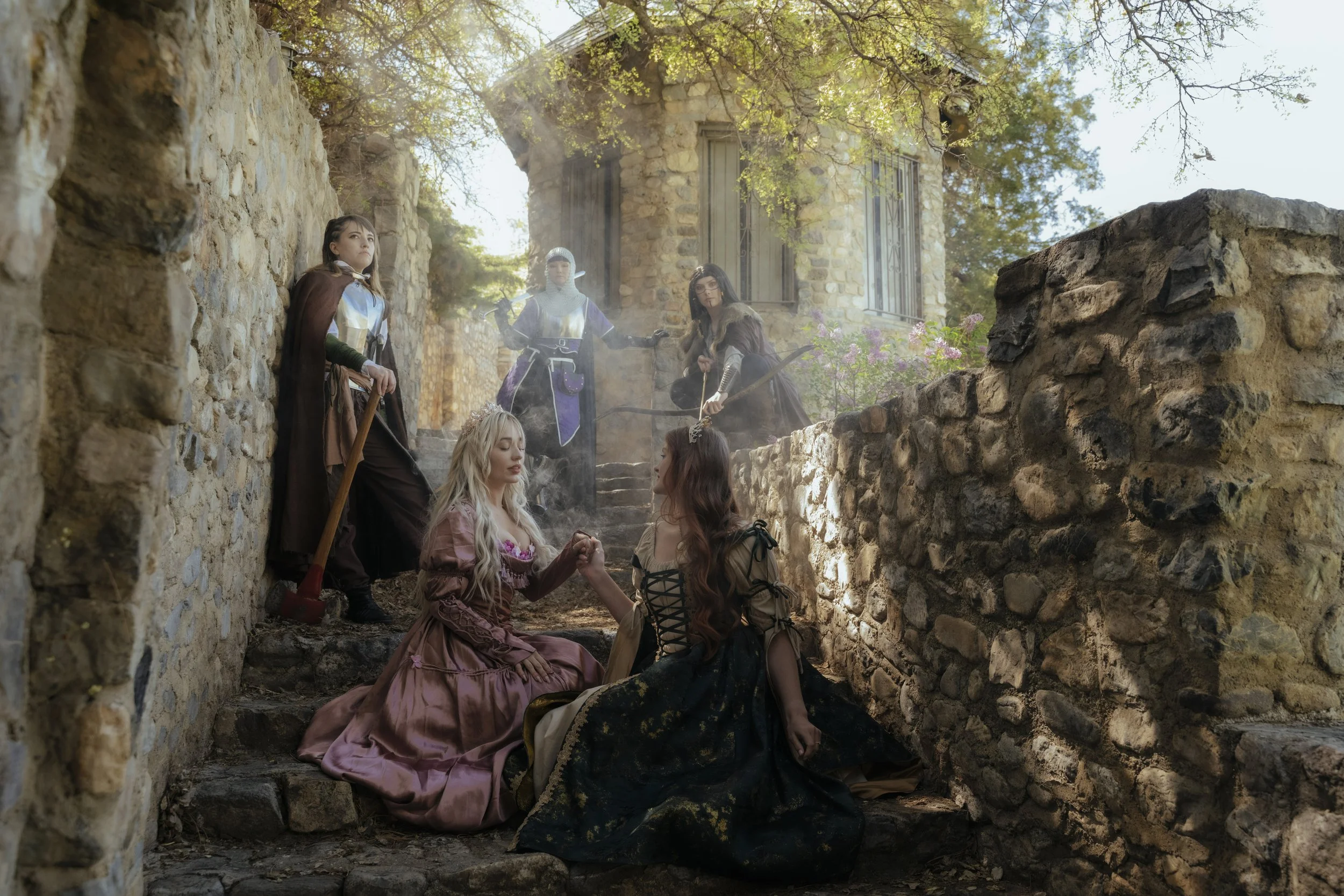Five women in medieval dresses and armor are on a stone staircase and courtyard, with one woman kneeling and holding hands with another, while the others stand around them, surrounded by stone walls and an old stone building with windows and trees.