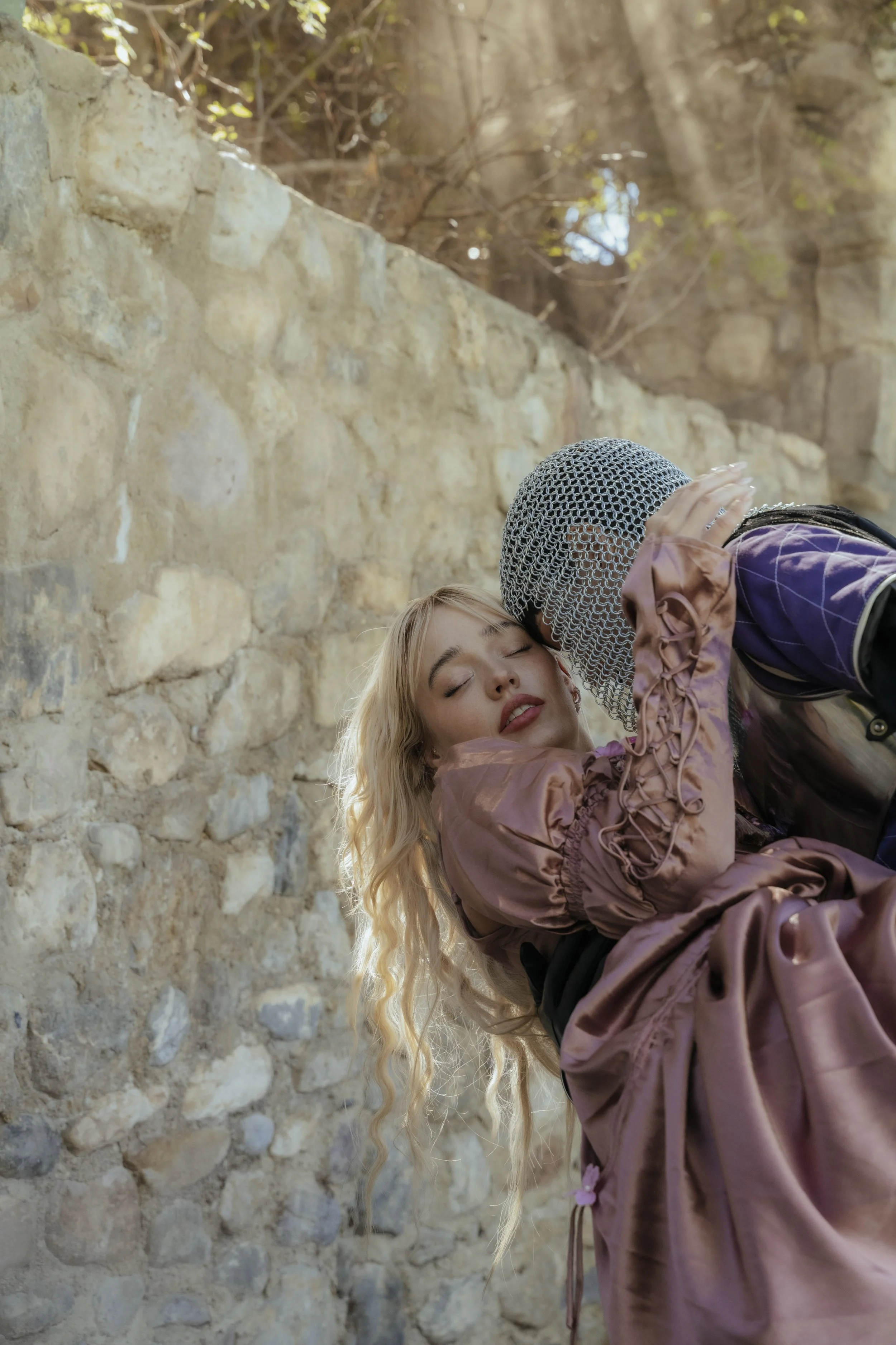 A woman with long blonde hair and a pink satin dress being kissed on the forehead by a person wearing medieval knight armor and chainmail, against a stone wall outdoor setting.