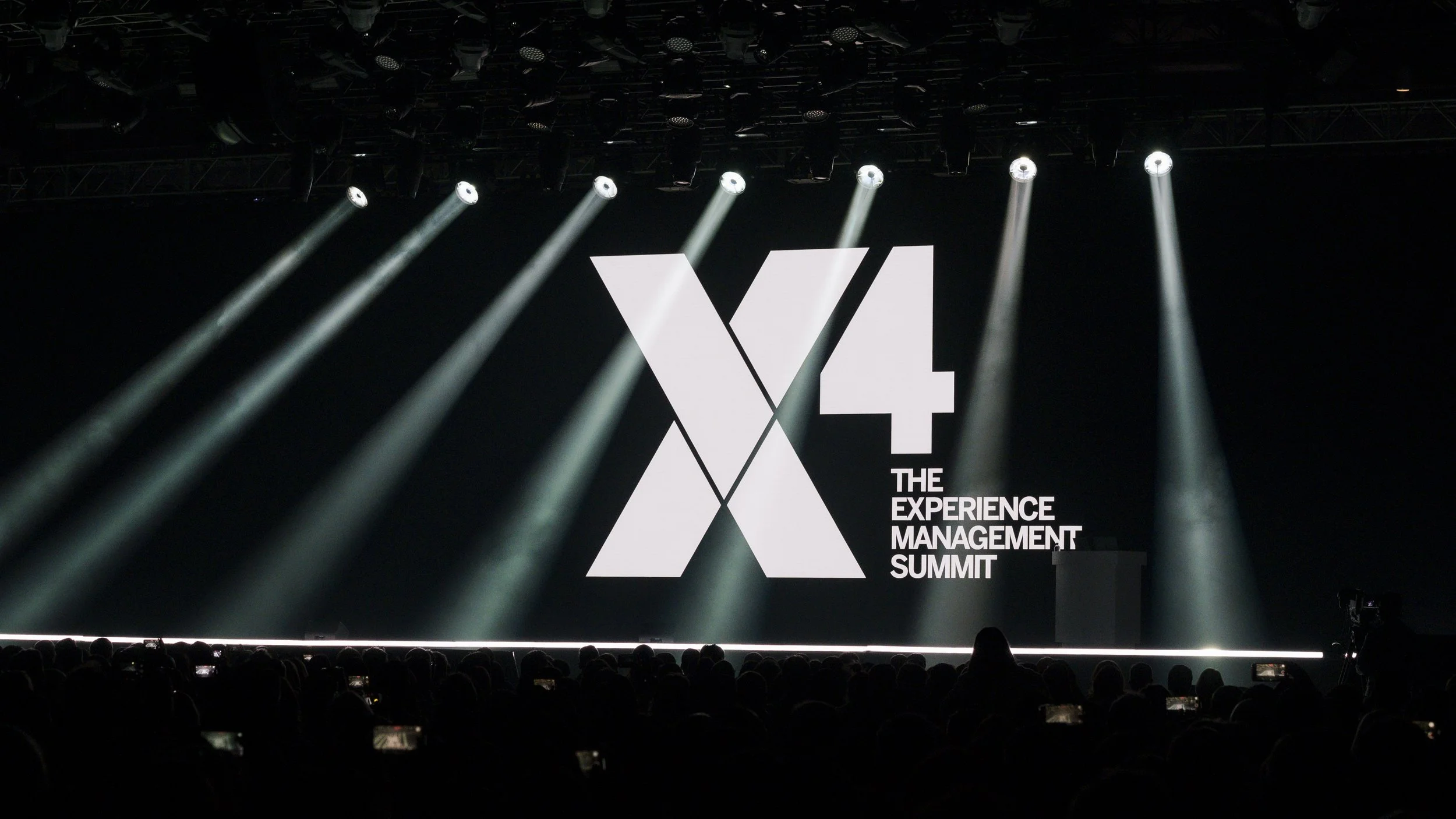 Stage with large screen displaying the logo of the X4 Experience Management Summit, with spotlights shining down and an audience in silhouette.