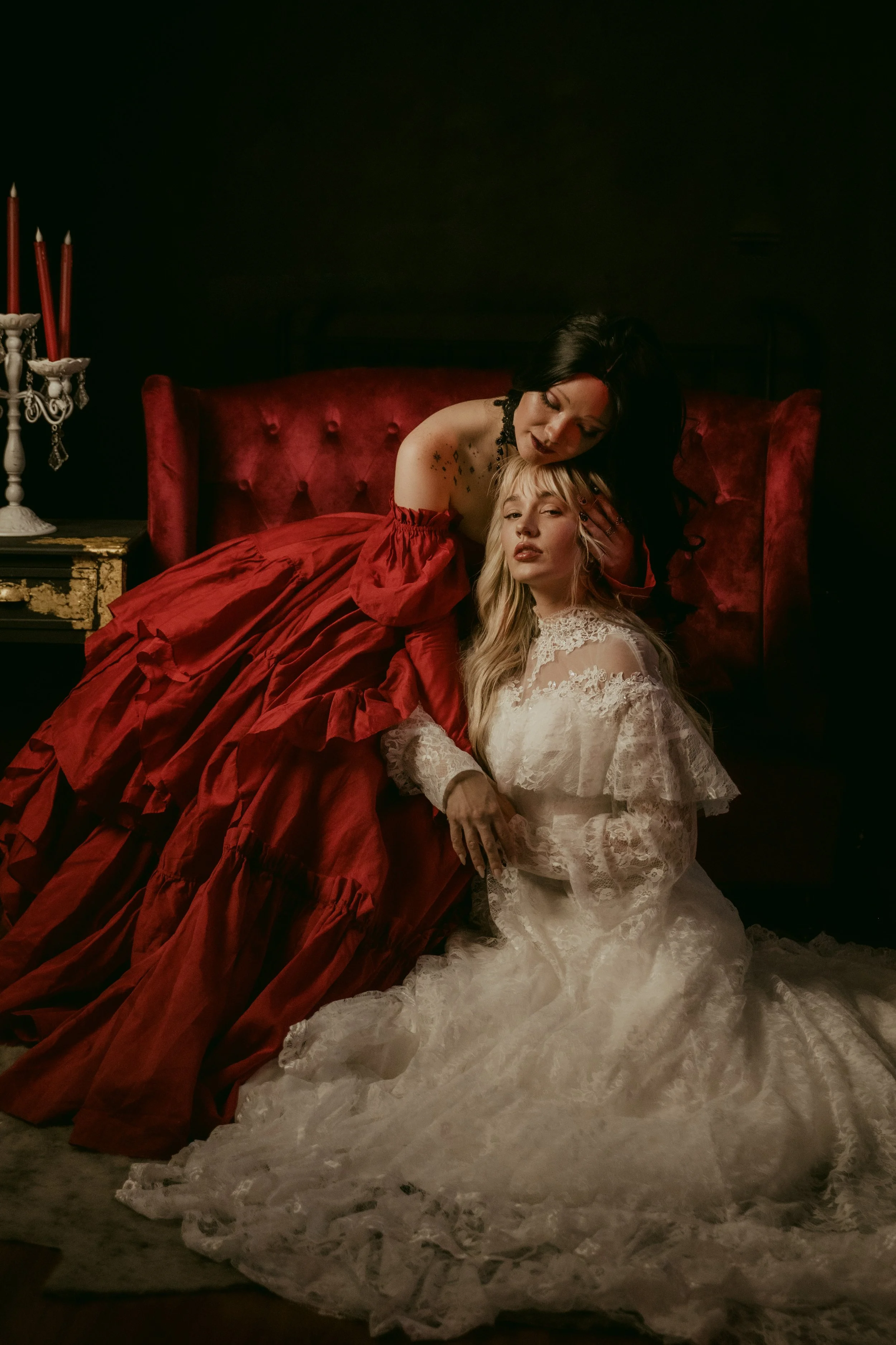Two women, one with black hair and dark makeup, wearing a red dress, and the other with blonde hair in a white lace dress, posing together in an elegant, vintage-inspired setting with a dark background and a candelabra on a table.
