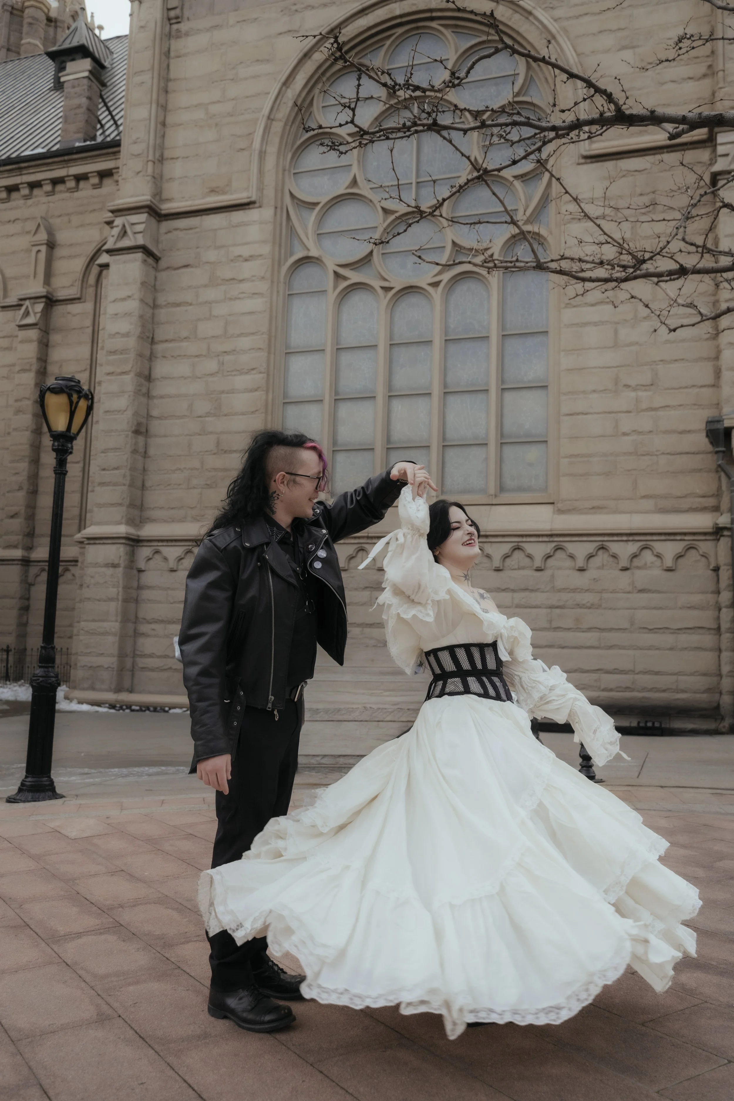 A person with a punk hairstyle and a leather jacket spins a woman in a vintage white dress outside a stone church or cathedral.