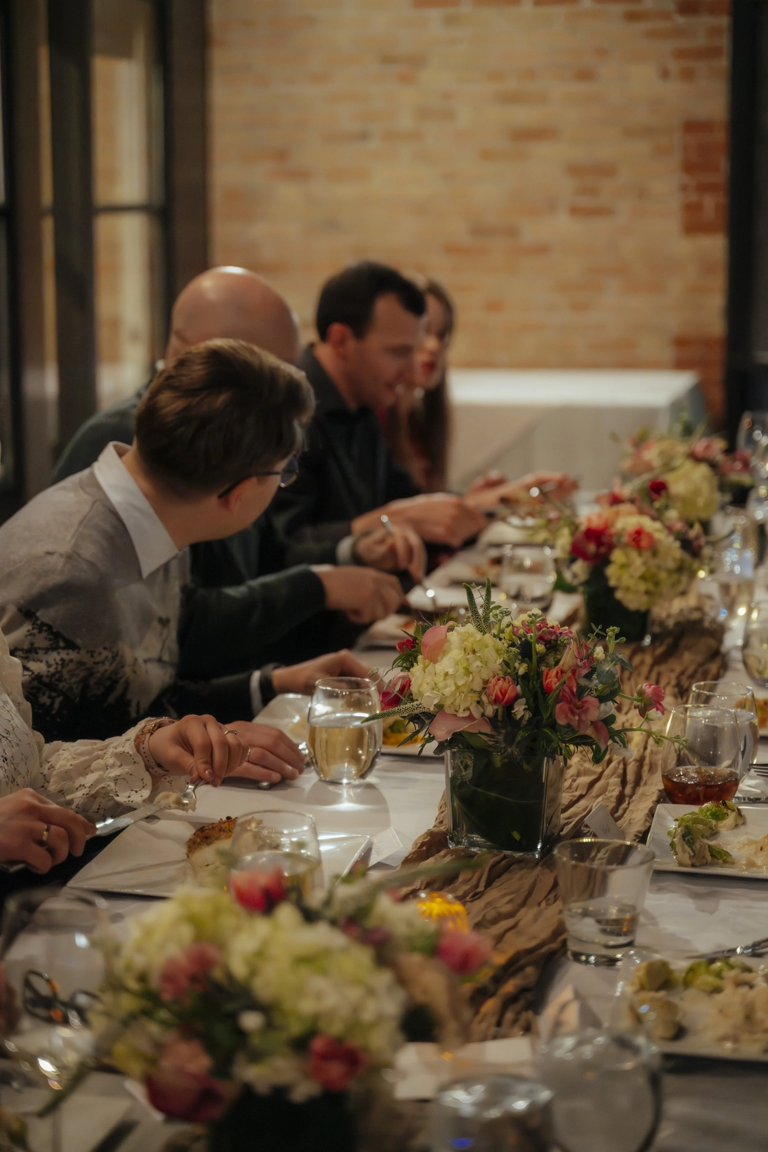 People sitting at a long dinner table with floral centerpieces, engaged in conversation, in a warm-lit indoor setting.