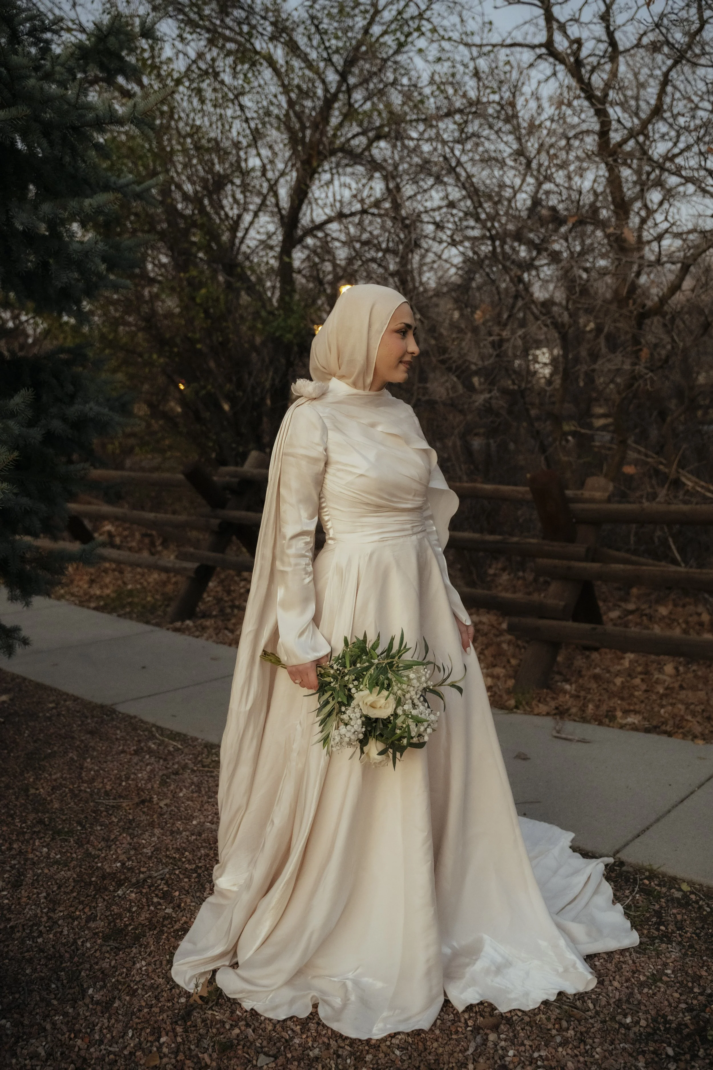 A woman in a cream-colored wedding gown and matching headscarf holding a bouquet of white roses and greenery, standing outdoors near trees with bare branches during dusk.