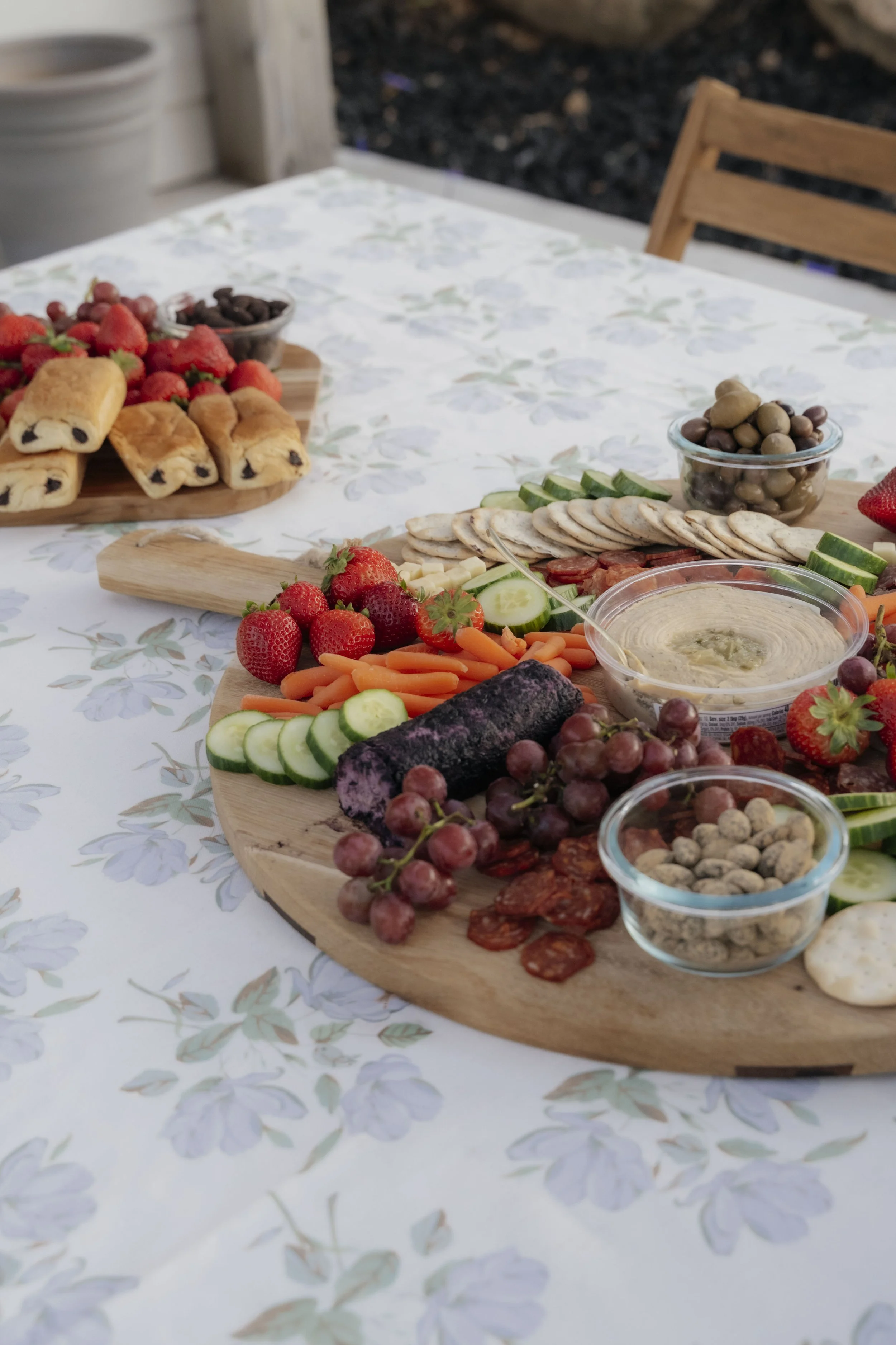 A charcuterie board with strawberries, grapes, cucumber, carrots, cucumber, sliced meats, crackers, dips, and olive bowls on a floral tablecloth, with additional strawberries, mini pastries, and olives in the background.