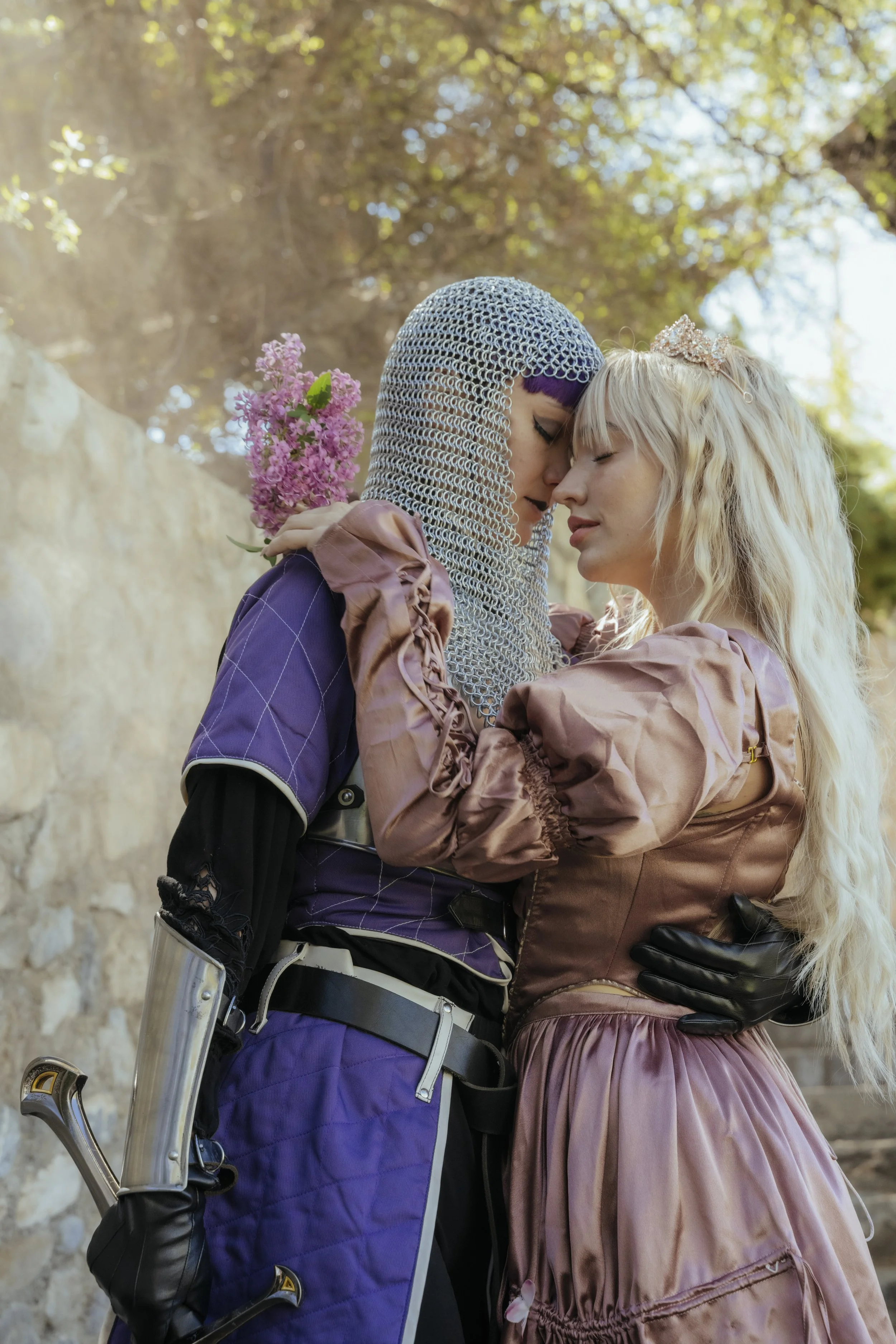Two women dressed in fantasy costumes embrace and touch foreheads outdoors, with trees and sunlight in the background.
