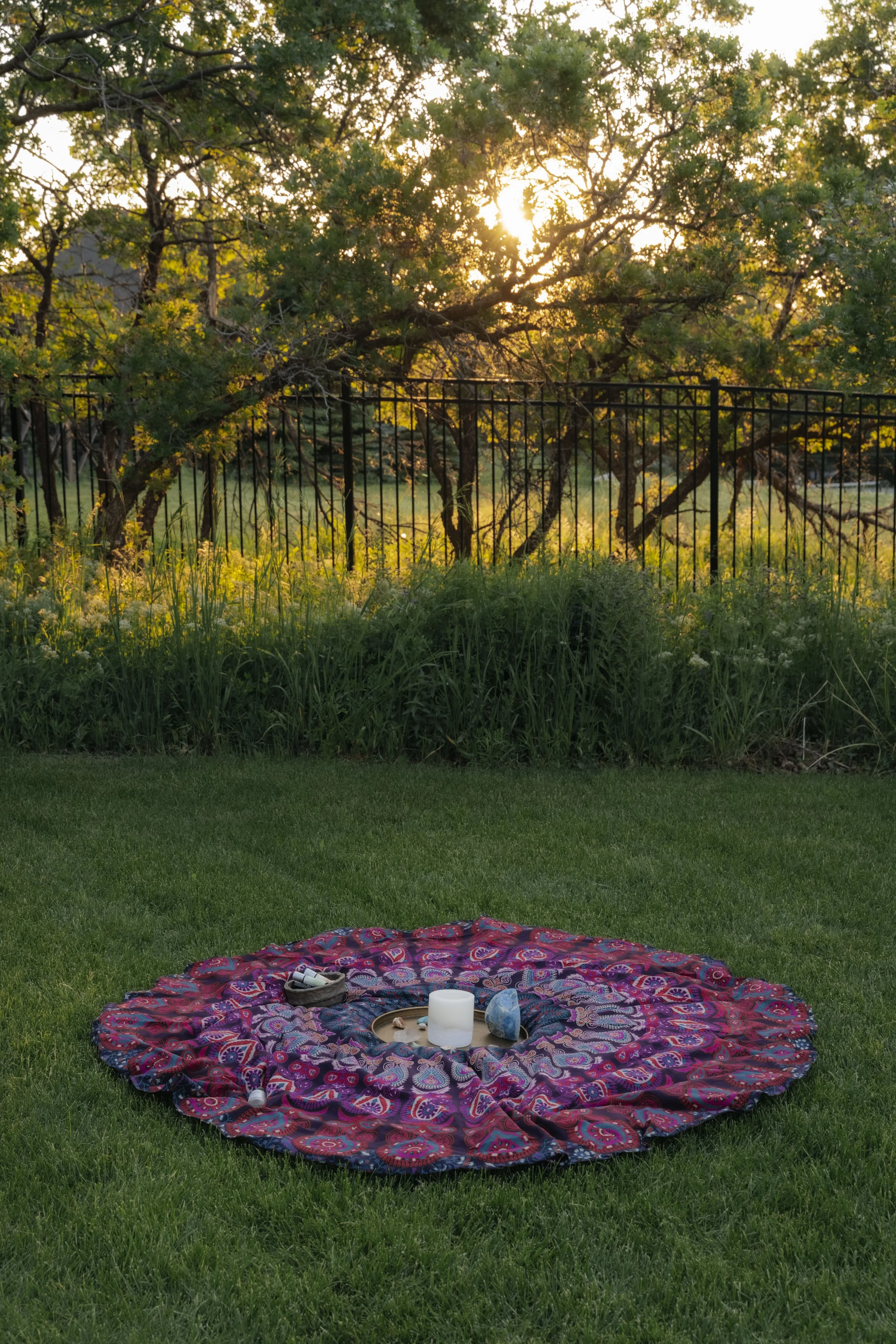 A round, colorful blanket with paisley patterns is spread on the grass with a candle, a blue crystal, a rock, and some small items arranged on it. In the background, a black metal fence separates the lawn from a garden with leafy trees and tall grass