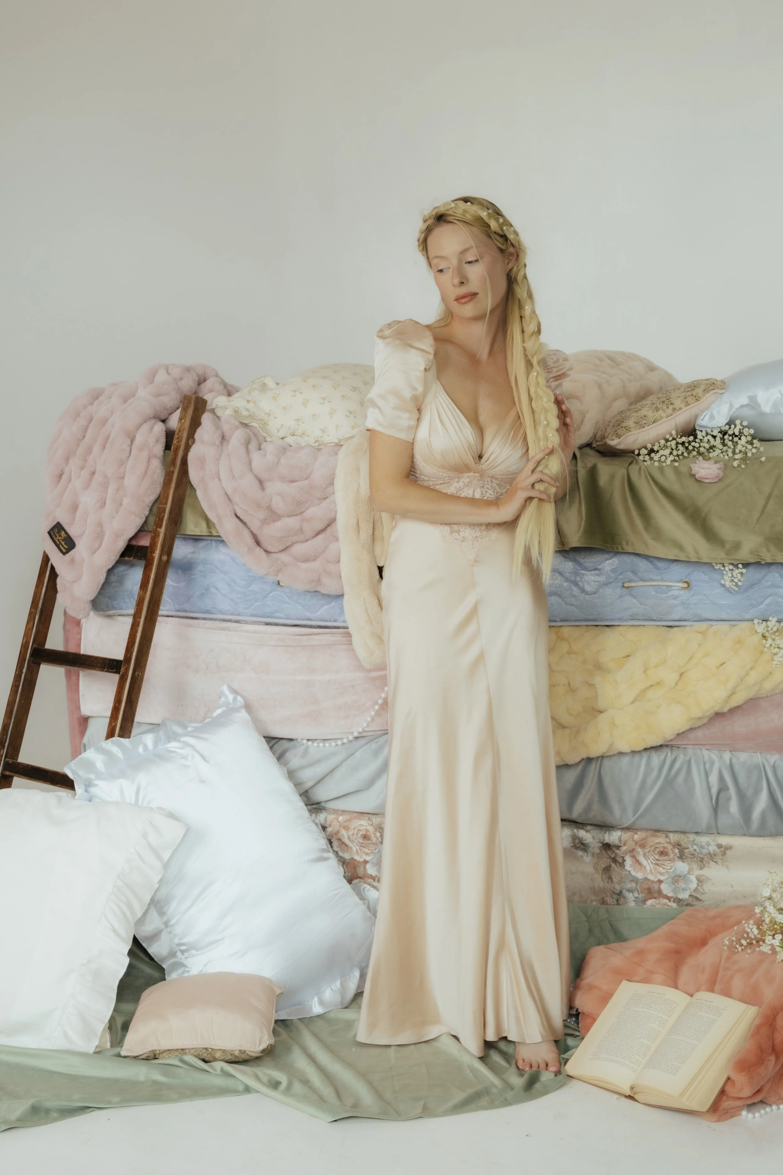 A woman with long braided blonde hair standing in front of a bed with various pillows and blankets, surrounded by books and fabric, in a minimalistic room with white walls.