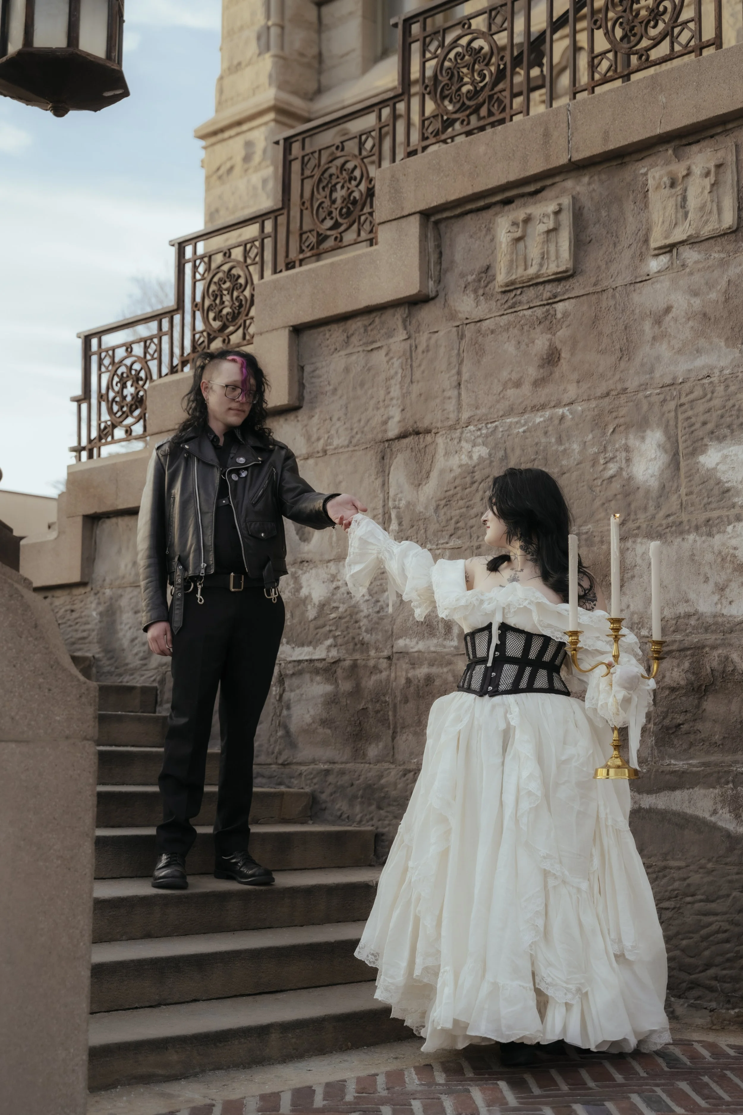 A person with black and pink hair, dressed in black leather clothing, is handing something to a woman in a white Victorian-style dress with a black corset. The woman has black hair and is holding a gold candelabrum with white candles. They are standi
