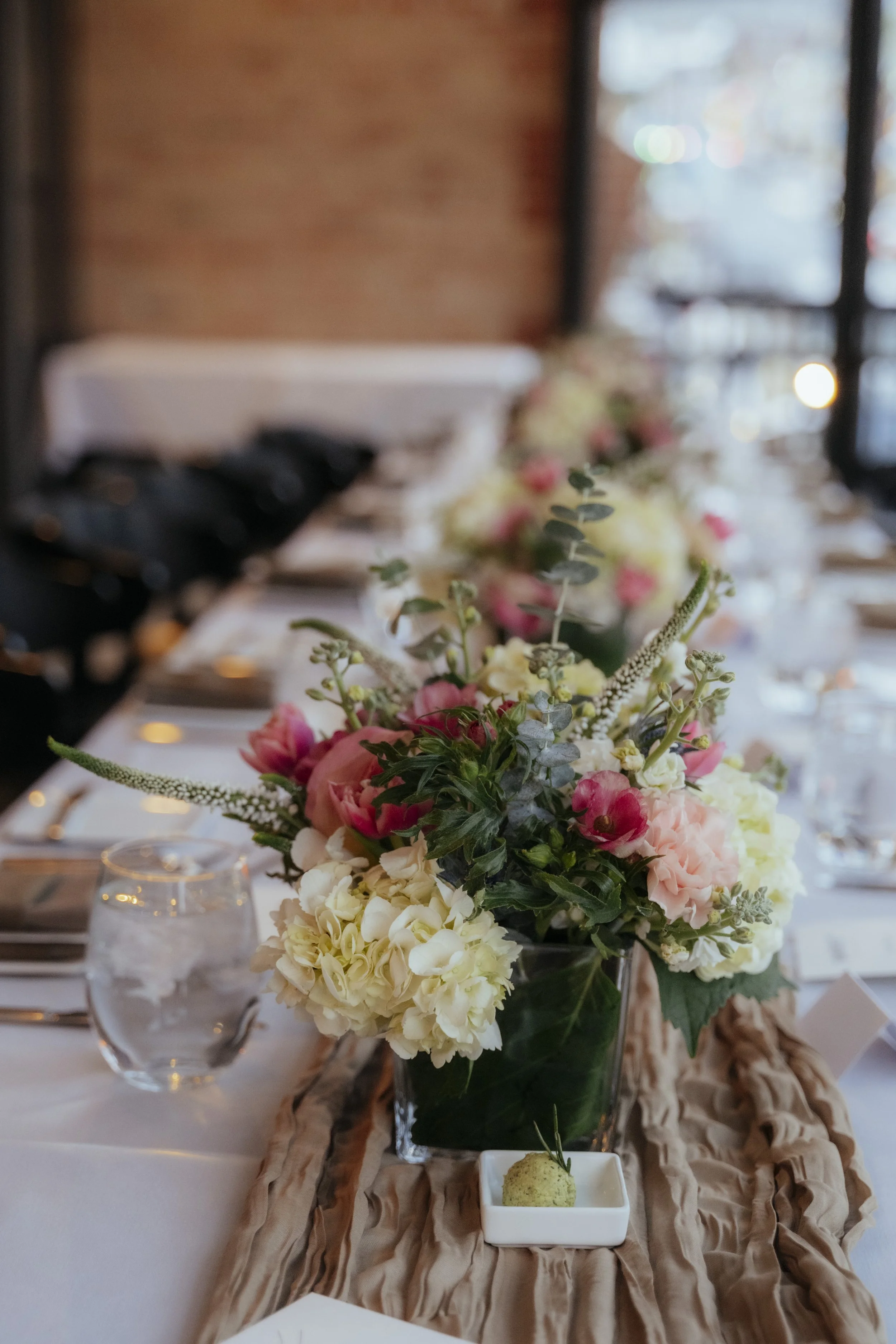 A floral centerpiece with pink, cream, and white flowers on a table, with a blurred view of a dining setup and windows in the background.